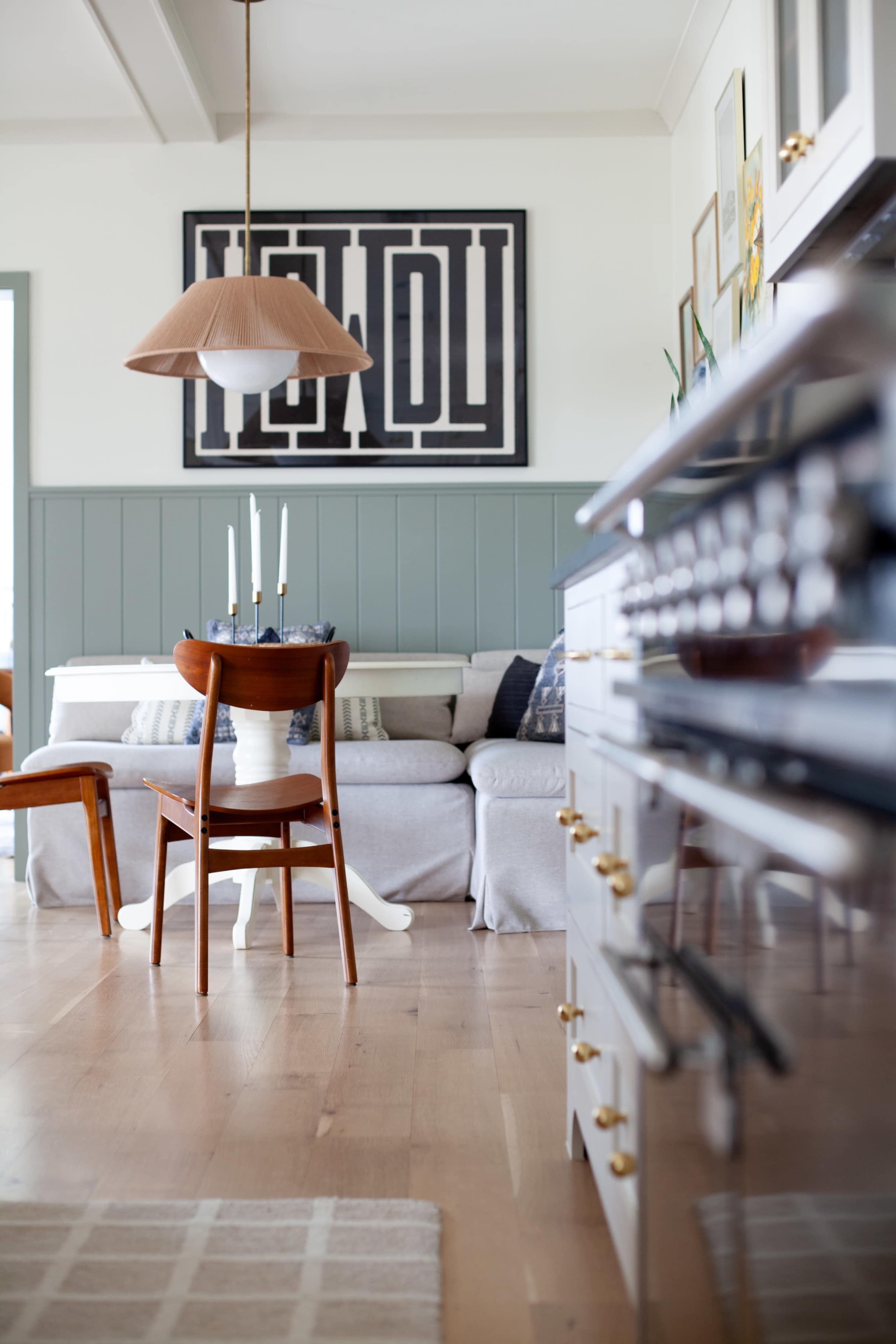 The image shows a modern kitchen and dining area with a gray sofa, wooden dining chairs, and a large piece of framed artwork on the wall.