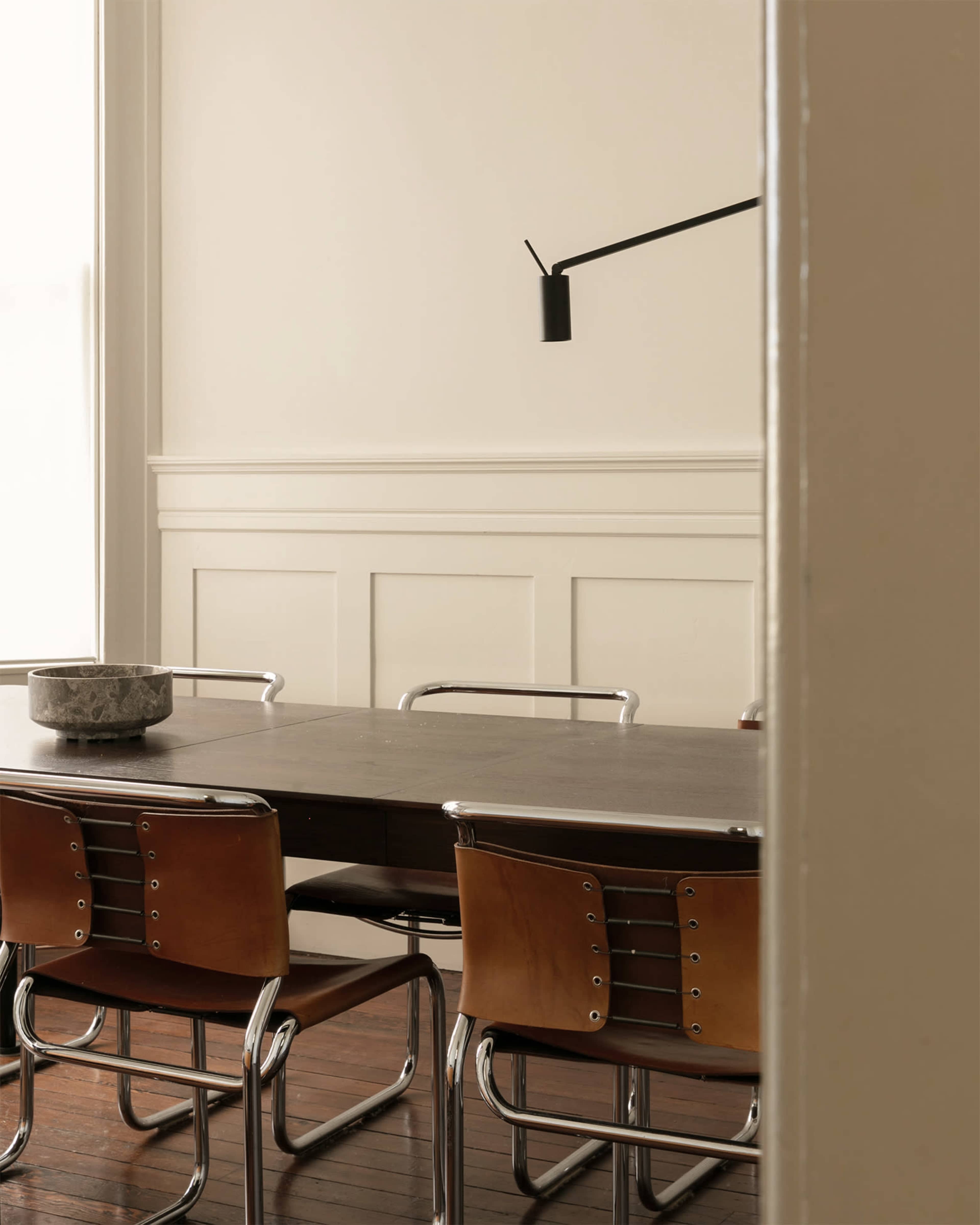 A wooden dining table is set in a room with a light wall, surrounded by several metal and leather chairs and illuminated by a wall-mounted lamp.