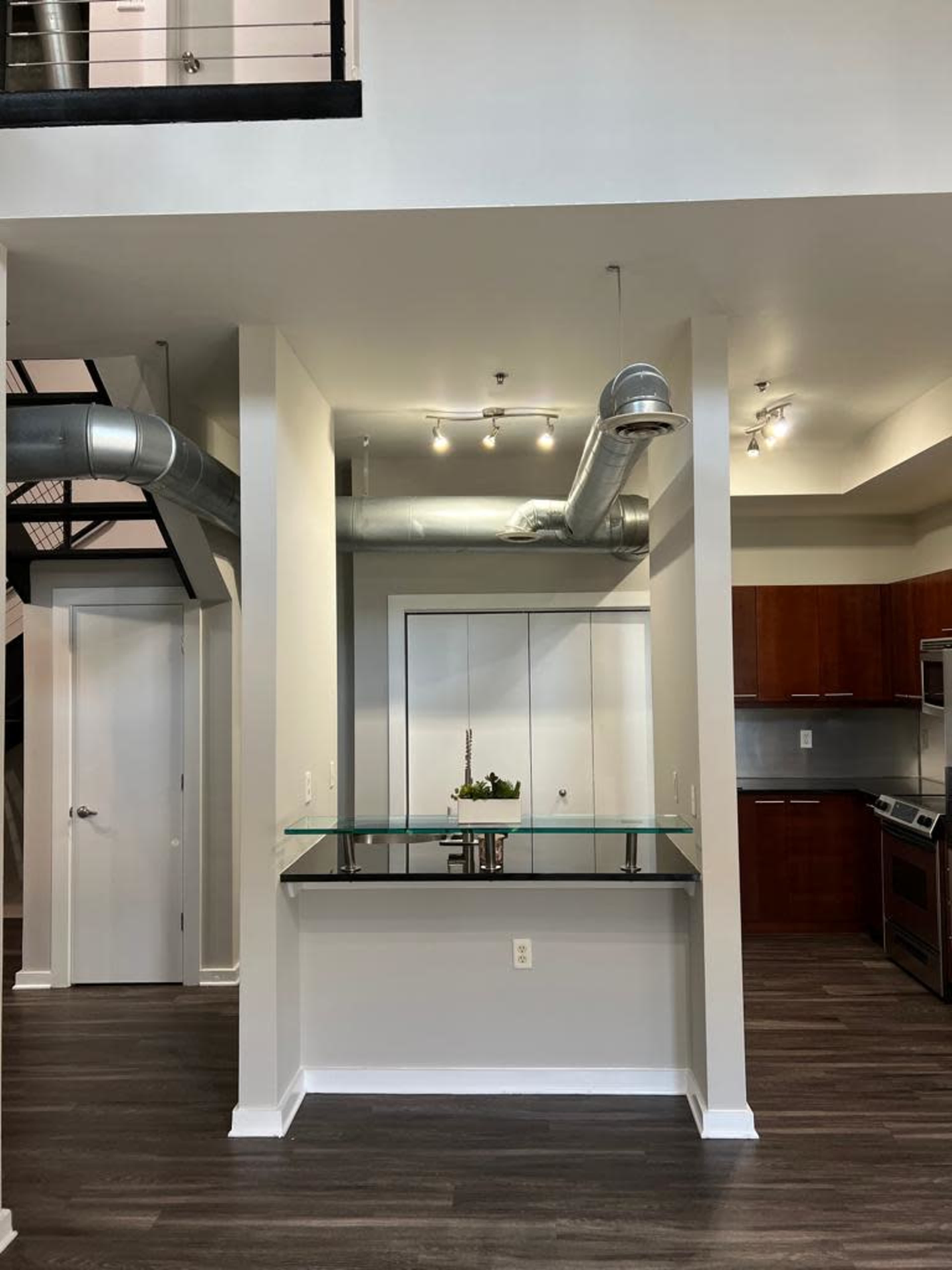 A modern kitchen area with a glass-top counter and stainless steel appliances, featuring exposed ductwork and a view of an adjacent room.