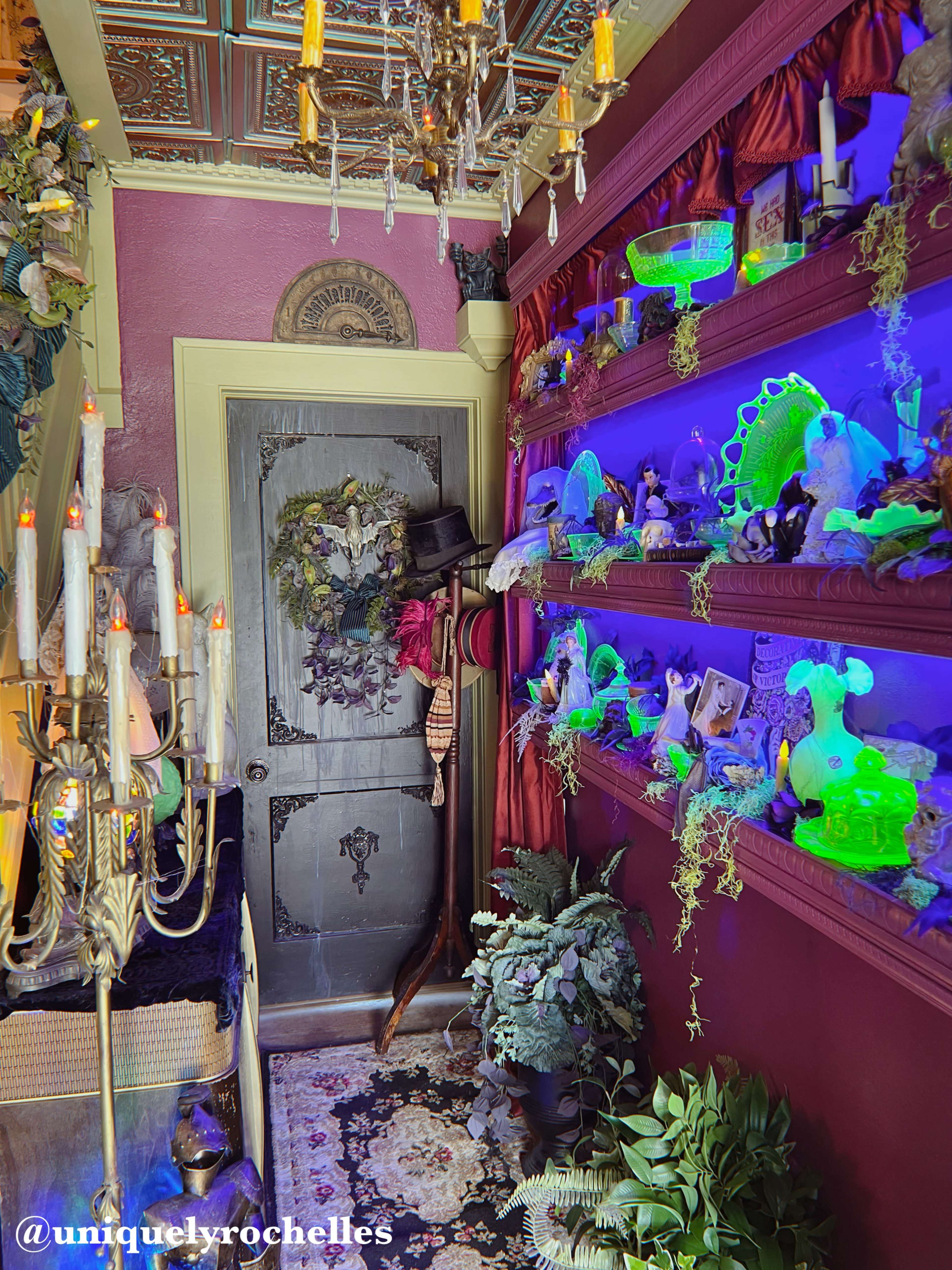 The image shows a narrow hallway decorated with a variety of colorful decorations, including illuminated shelves filled with figurines and plants, along with a vintage door adorned with a wreath.