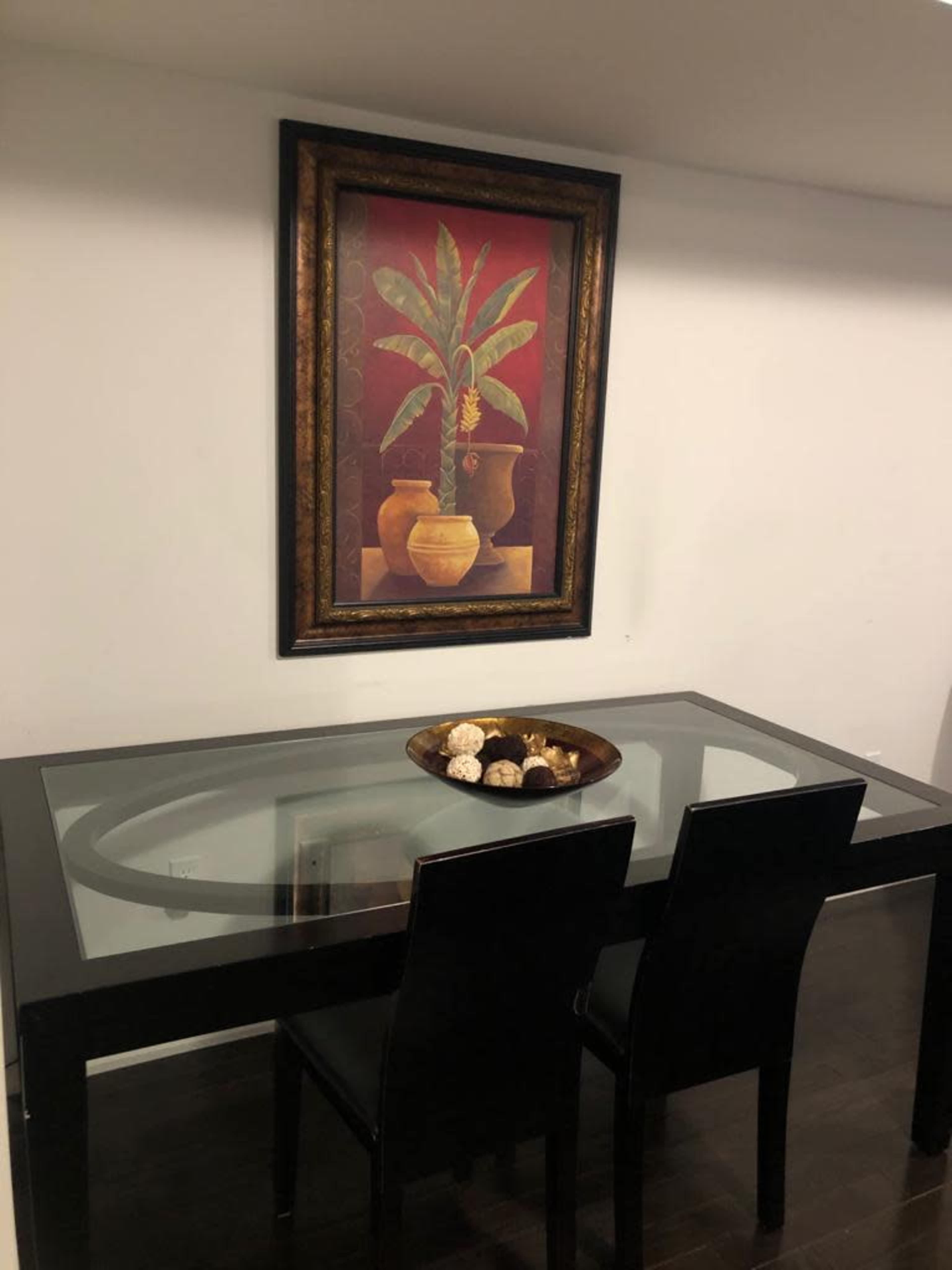 A glass-topped table with two black chairs sits beneath a framed painting of a plant and vases on a white wall.