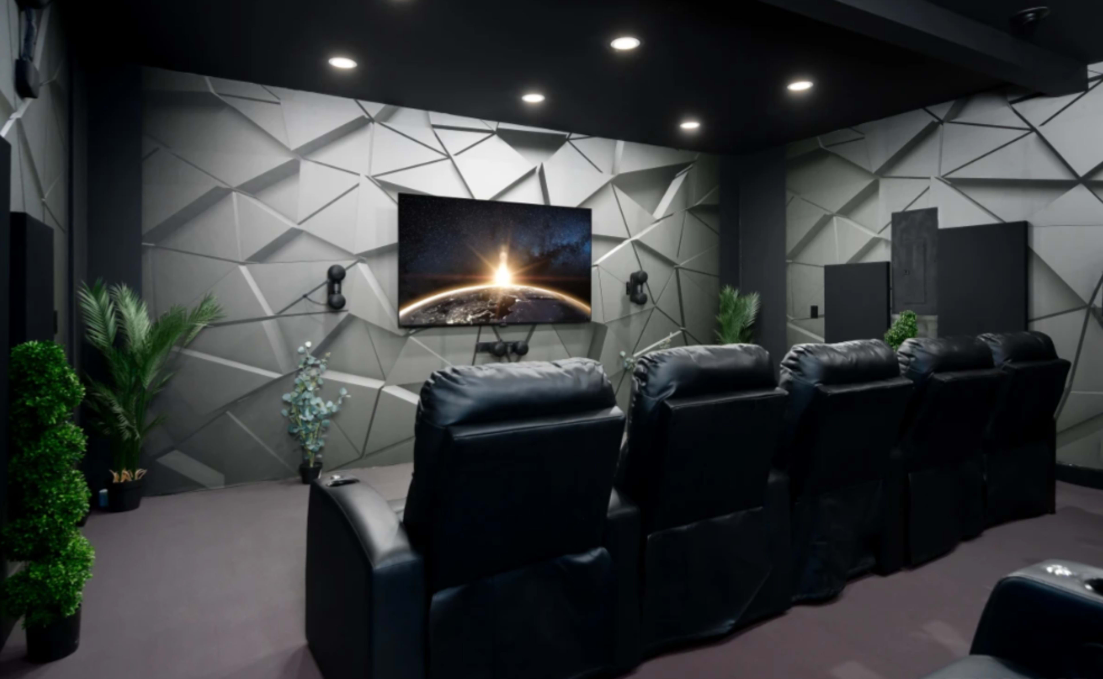 A modern home theater with black recliner seats facing a large wall-mounted screen, framed by geometric gray walls and decorative plants.