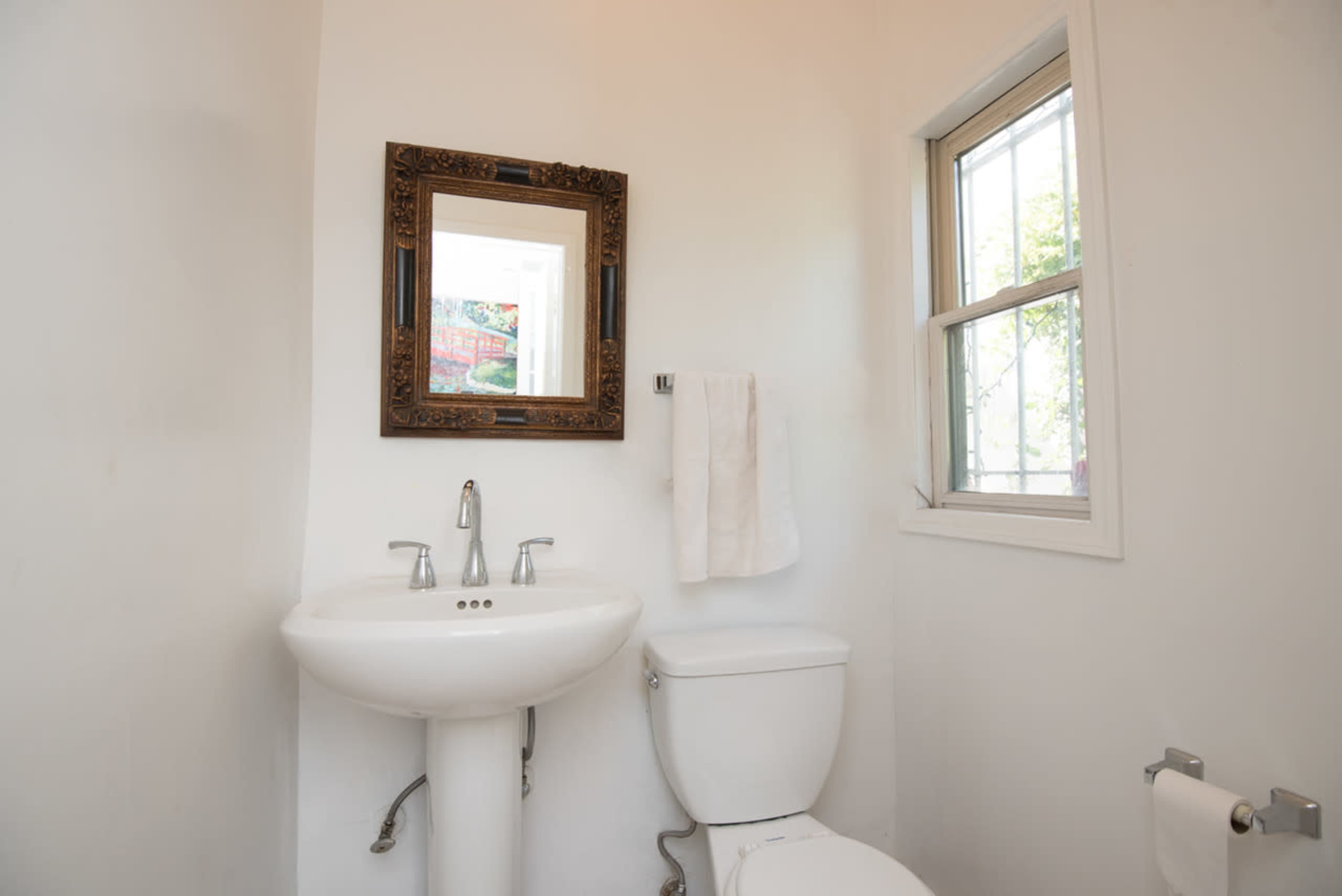 The image shows a small bathroom with a pedestal sink, a toilet, a mirror, and a window.