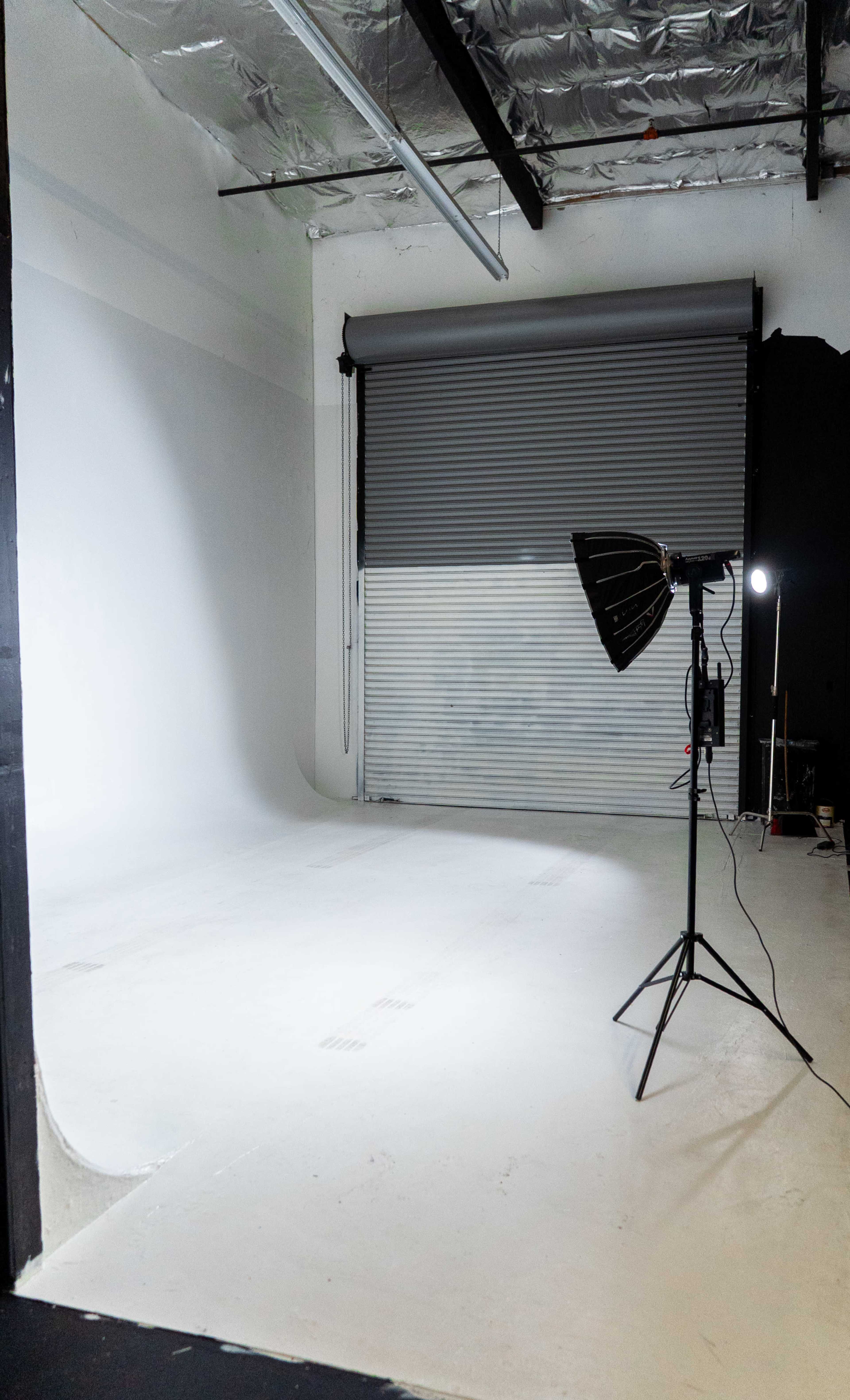 The image shows a photography studio featuring a seamless backdrop, a softbox light on a stand, and a large window with a roller shade.
