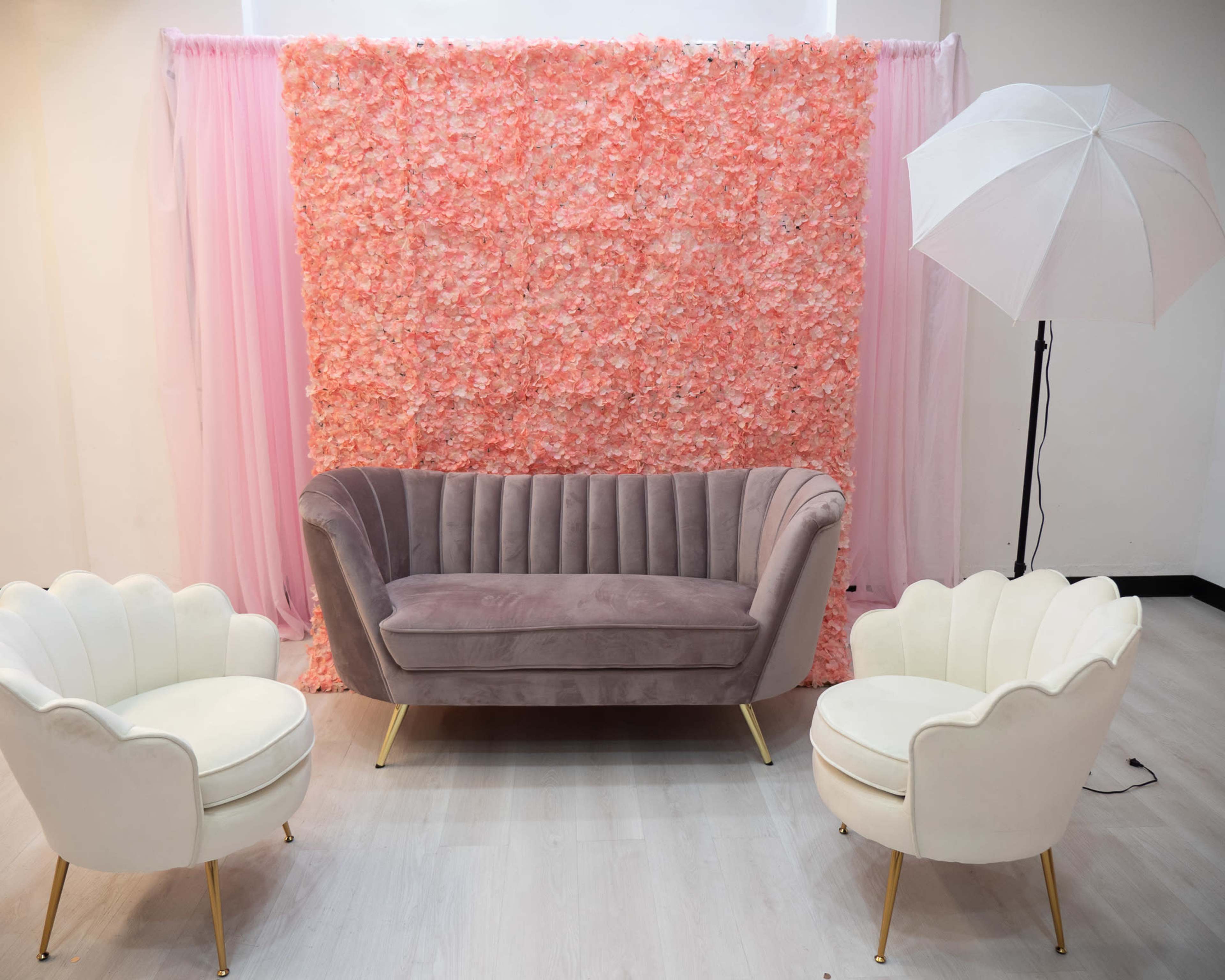 A stylish seating area with a grey velvet couch and two white chairs, set against a backdrop of pink floral decorations.