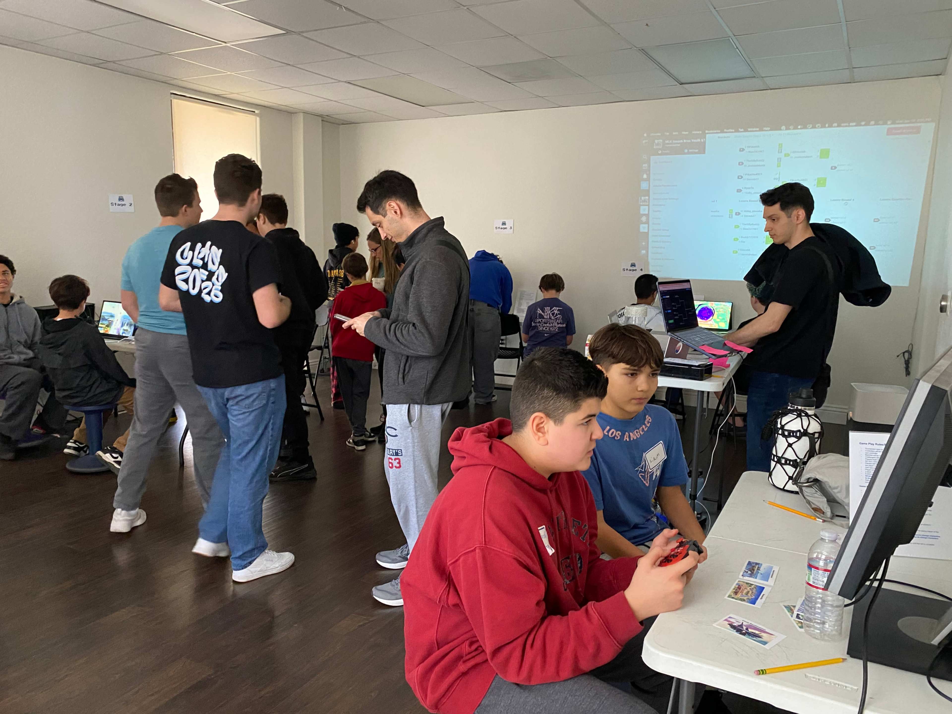 A group of students is gathered in a classroom, some working on computers while others interact with each other.