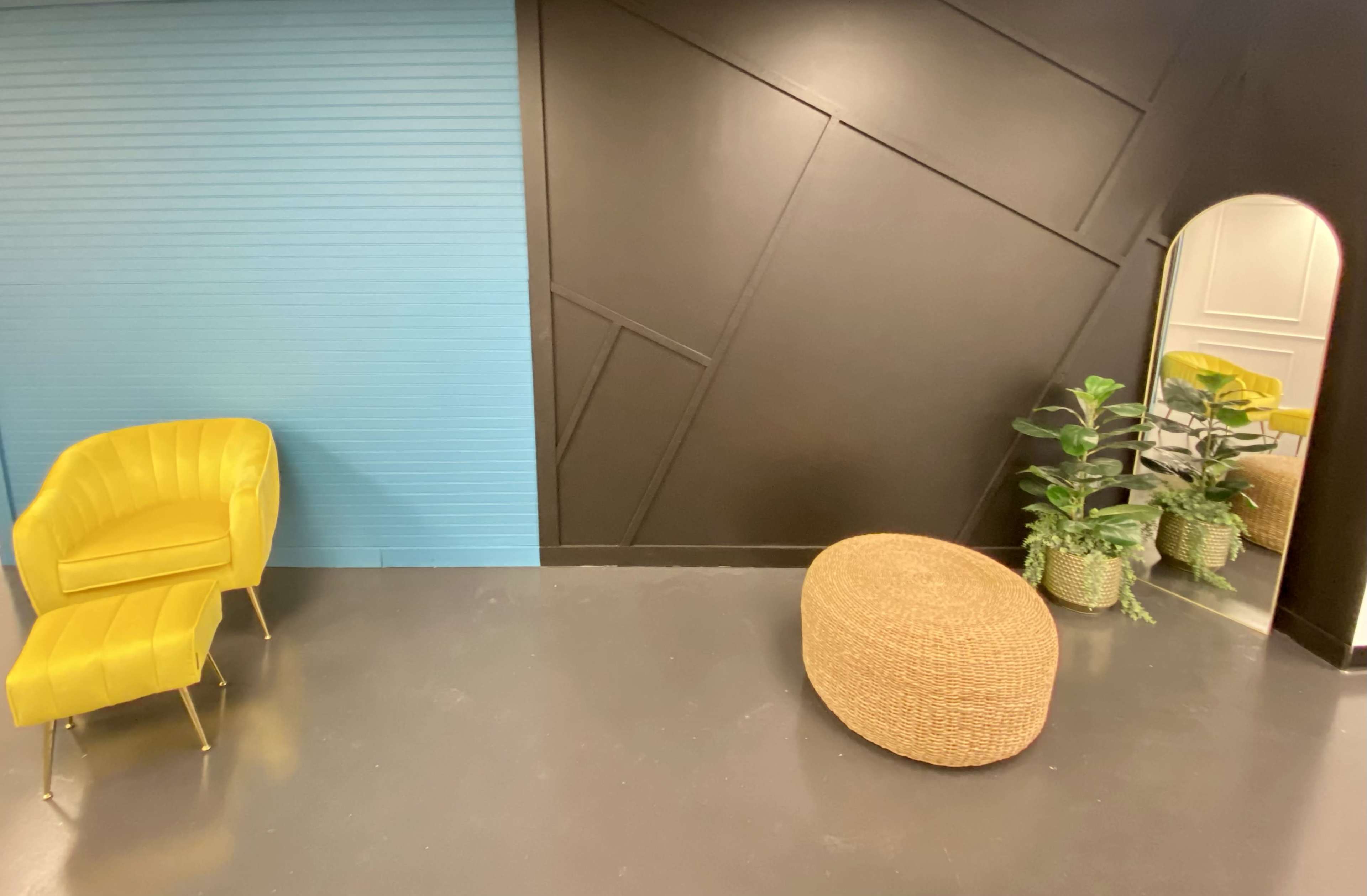 The image features a bright yellow chair and a round wicker ottoman, positioned near a wall with a blue section and a mirror, alongside two potted plants.