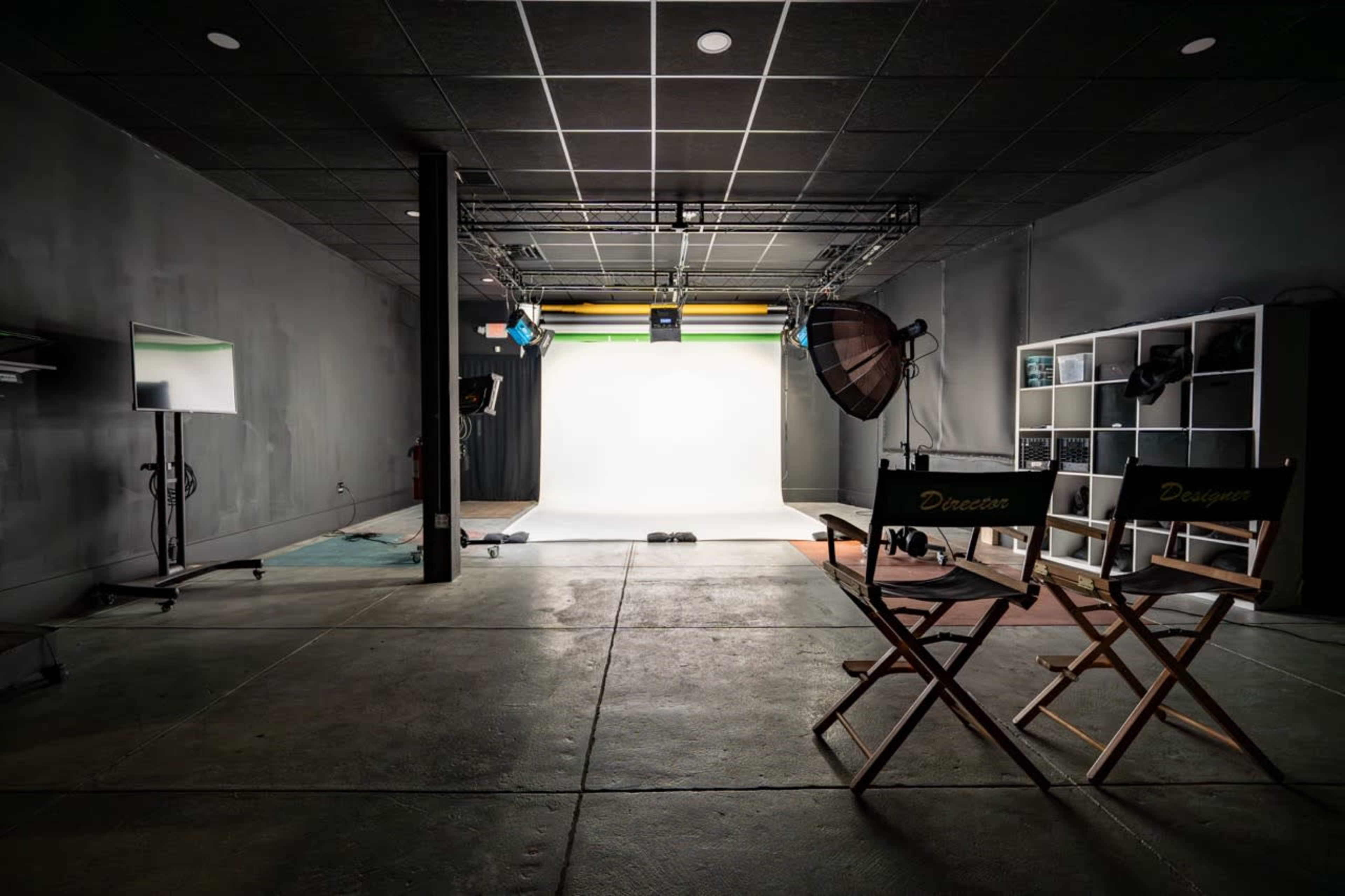 The image shows a spacious photography studio with two director's chairs facing a lit backdrop, equipped with various lighting and camera equipment.