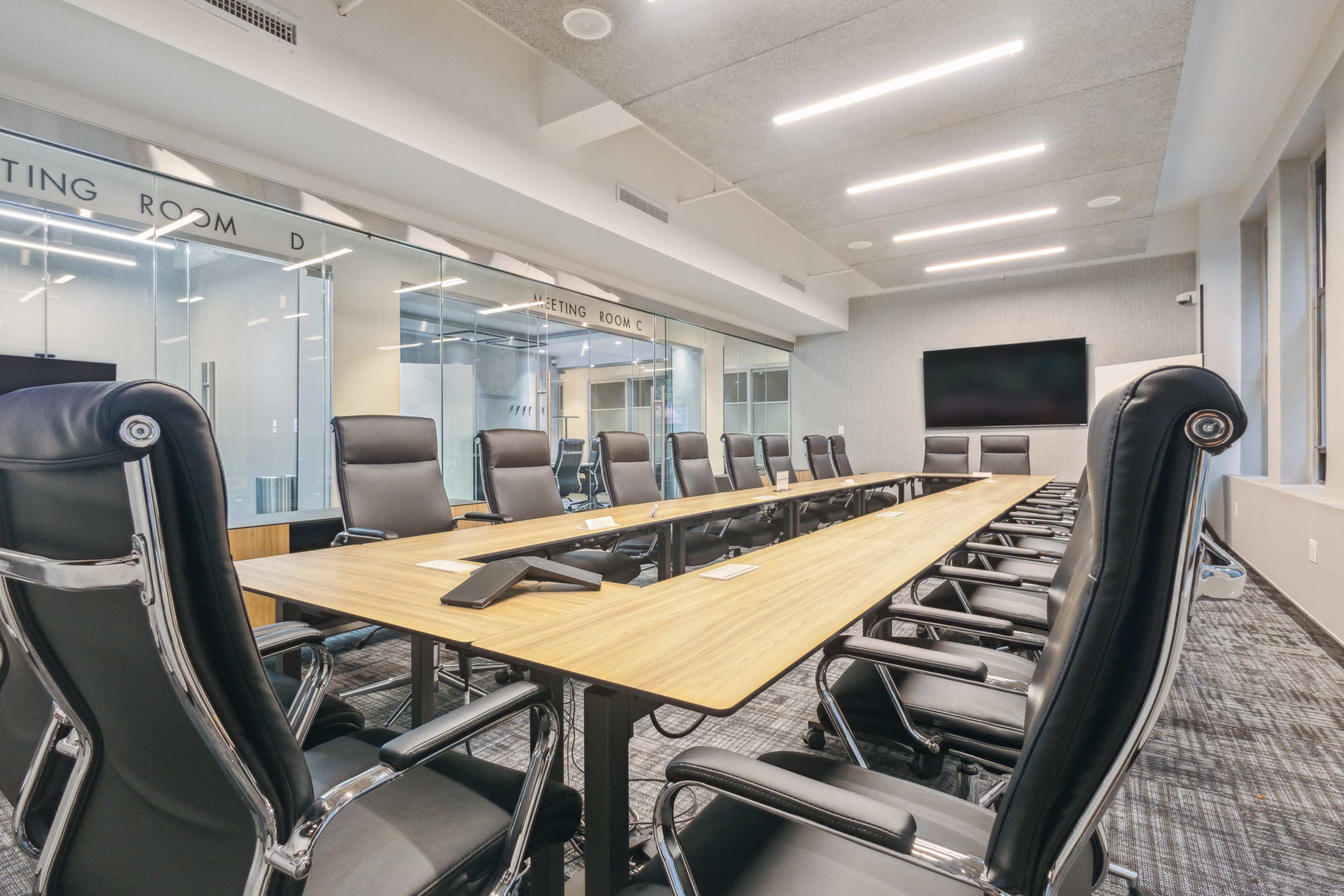 A long wooden conference table with multiple black office chairs is positioned in a modern meeting room featuring large windows and a wall-mounted screen.