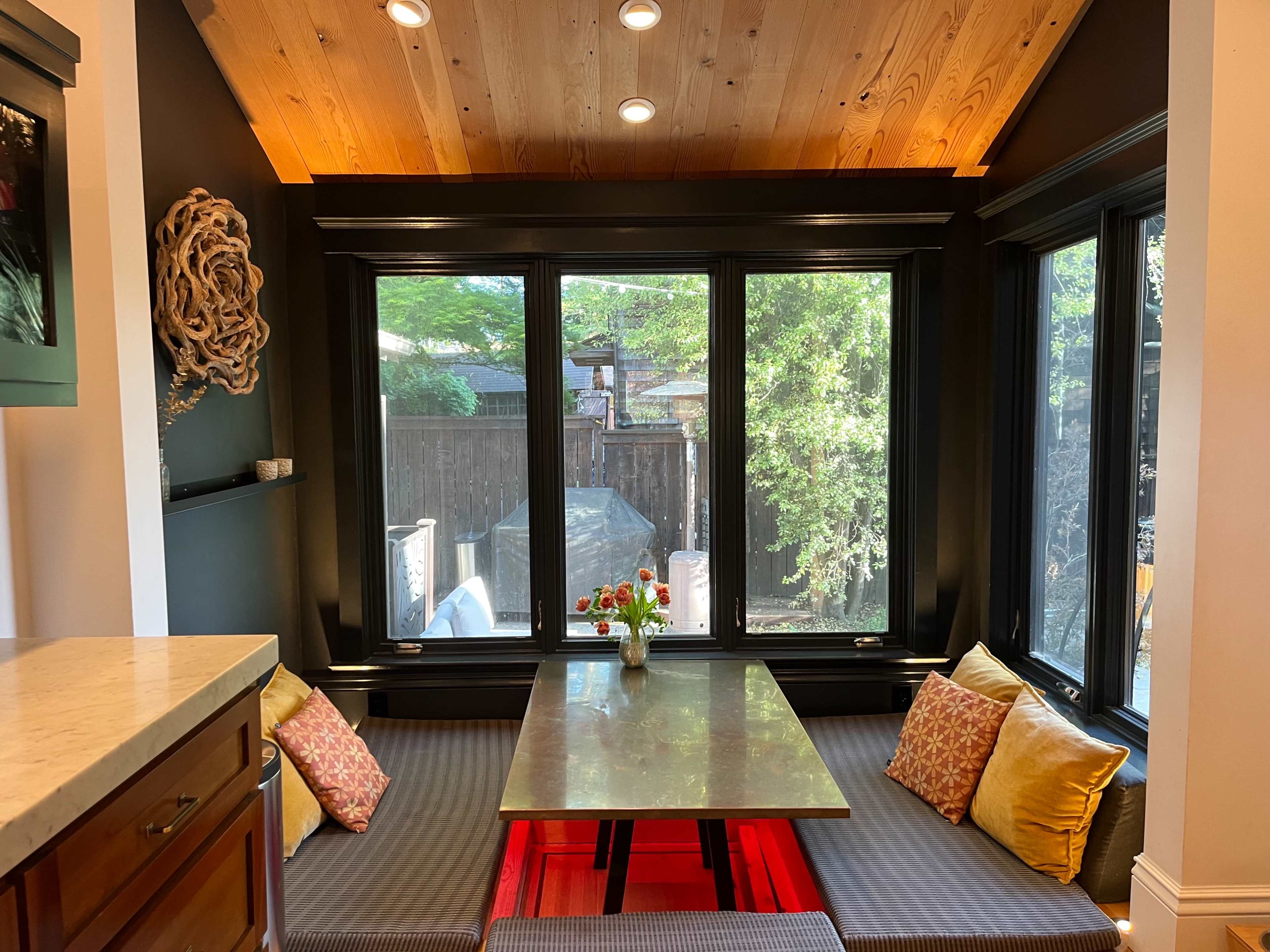 A cozy dining nook with a wooden table surrounded by cushioned bench seating, situated next to large windows that offer a view of greenery outside.