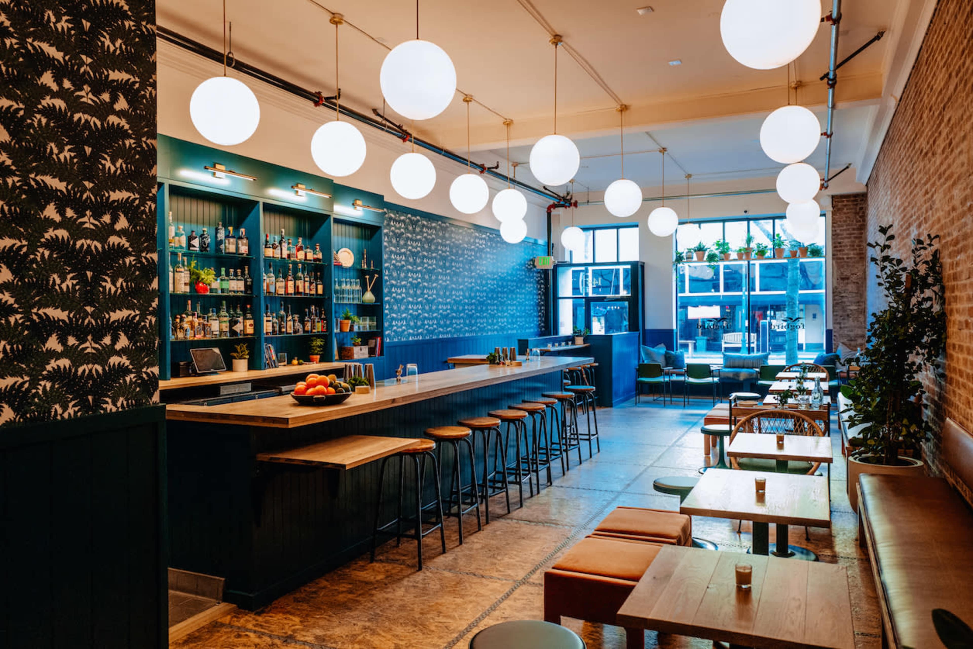 A modern bar with a long countertop, high stools, and large round lights hanging from the ceiling, surrounded by wooden tables and chairs.