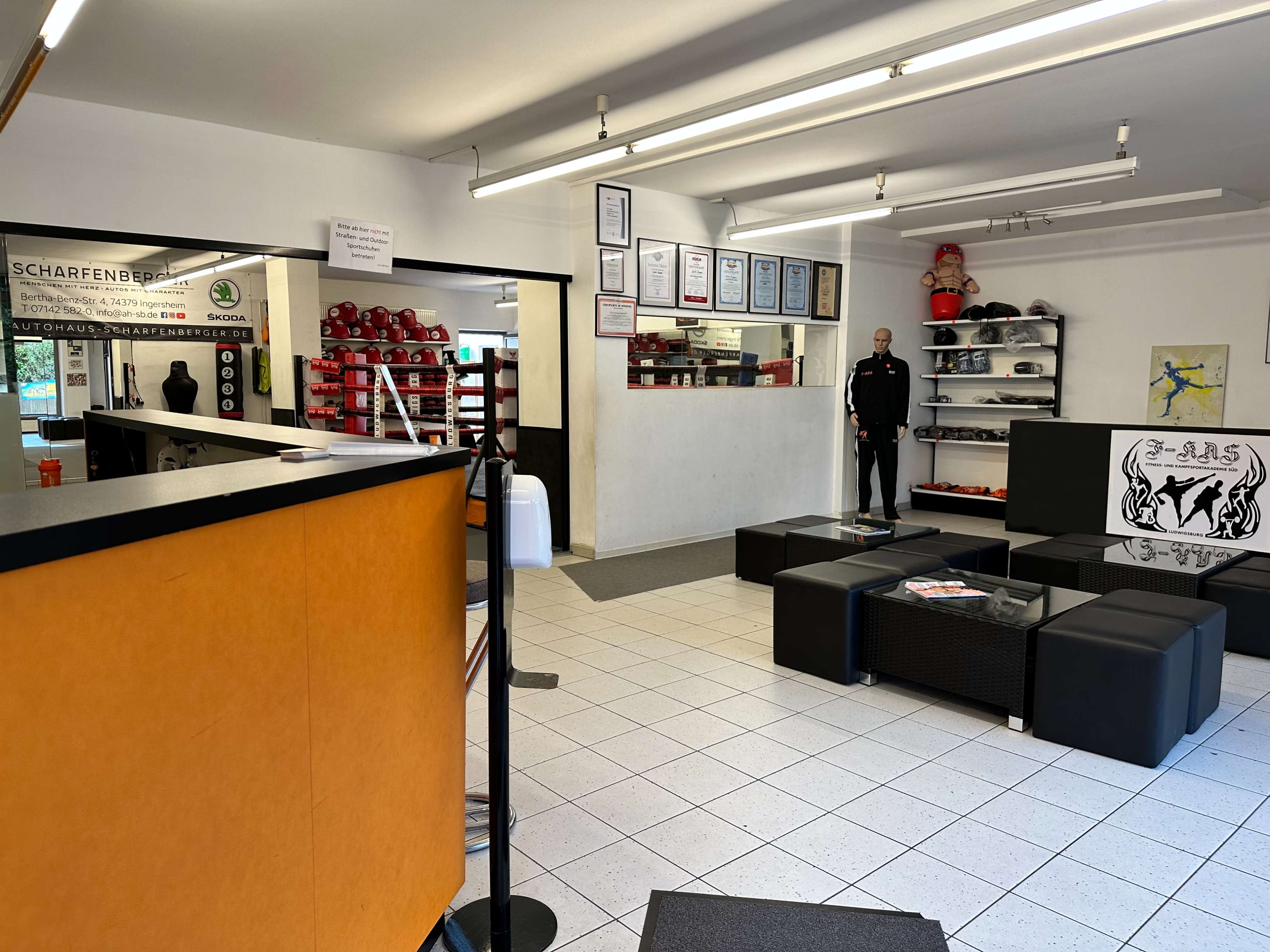 The image shows an indoor space with a reception area, featuring an orange counter, black seating, and a wall displaying various certificates and boxing equipment.
