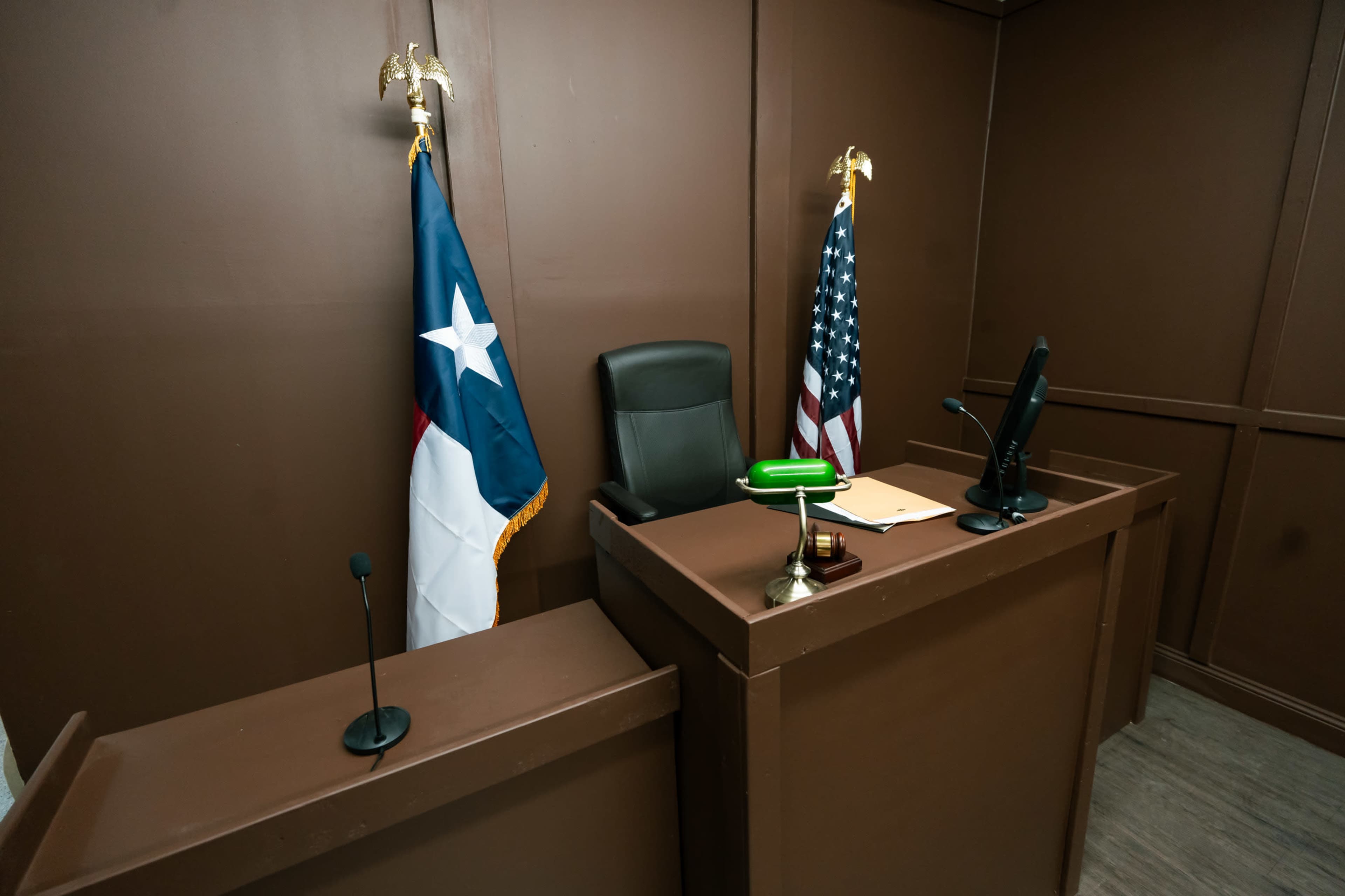 A courtroom scene featuring a Texas state flag and an American flag flanking a judge's bench with a black chair and a green gavel.