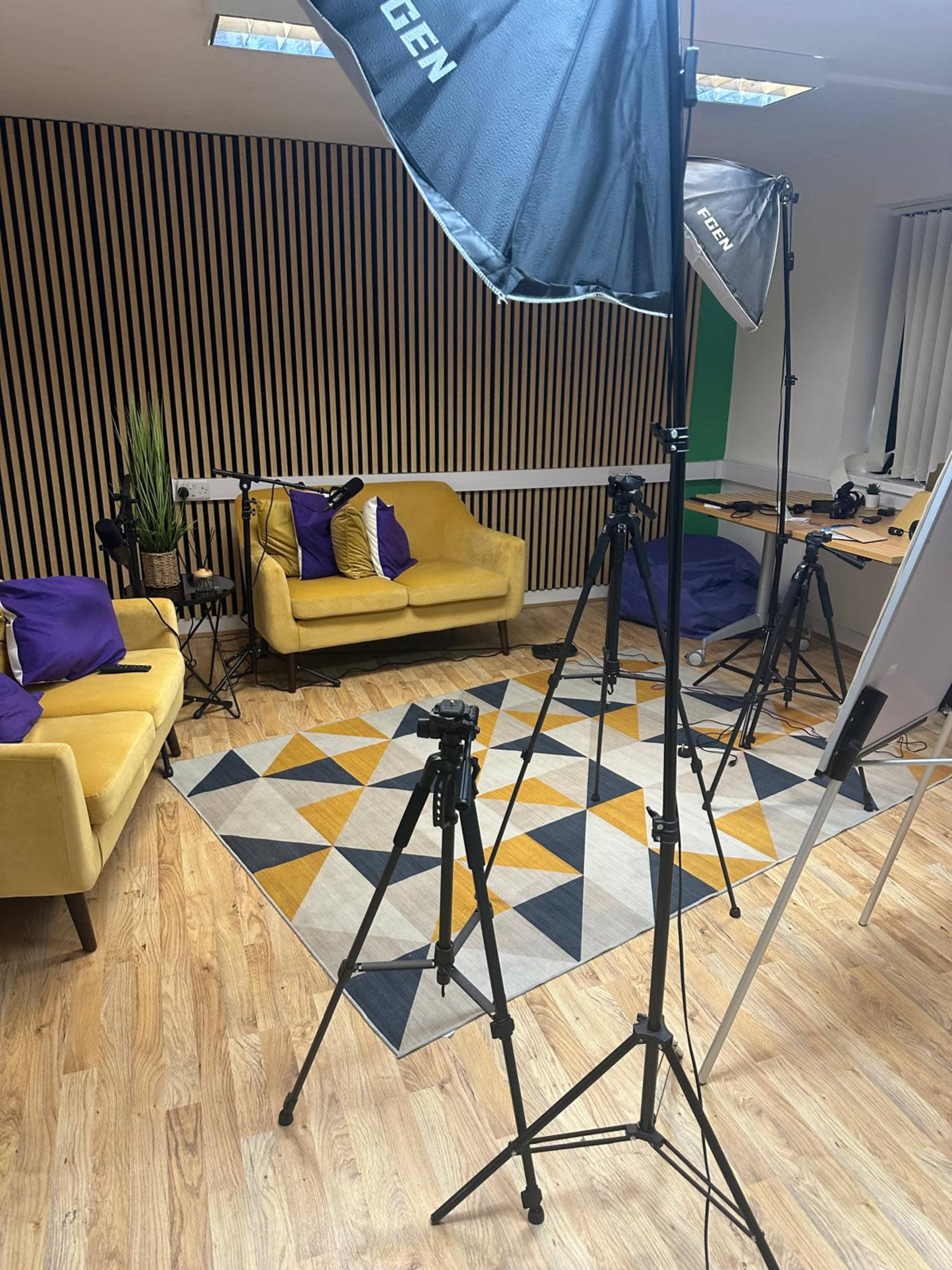 The image shows a studio setup with yellow sofas, a geometric rug, and several tripods and lighting equipment arranged around the space.