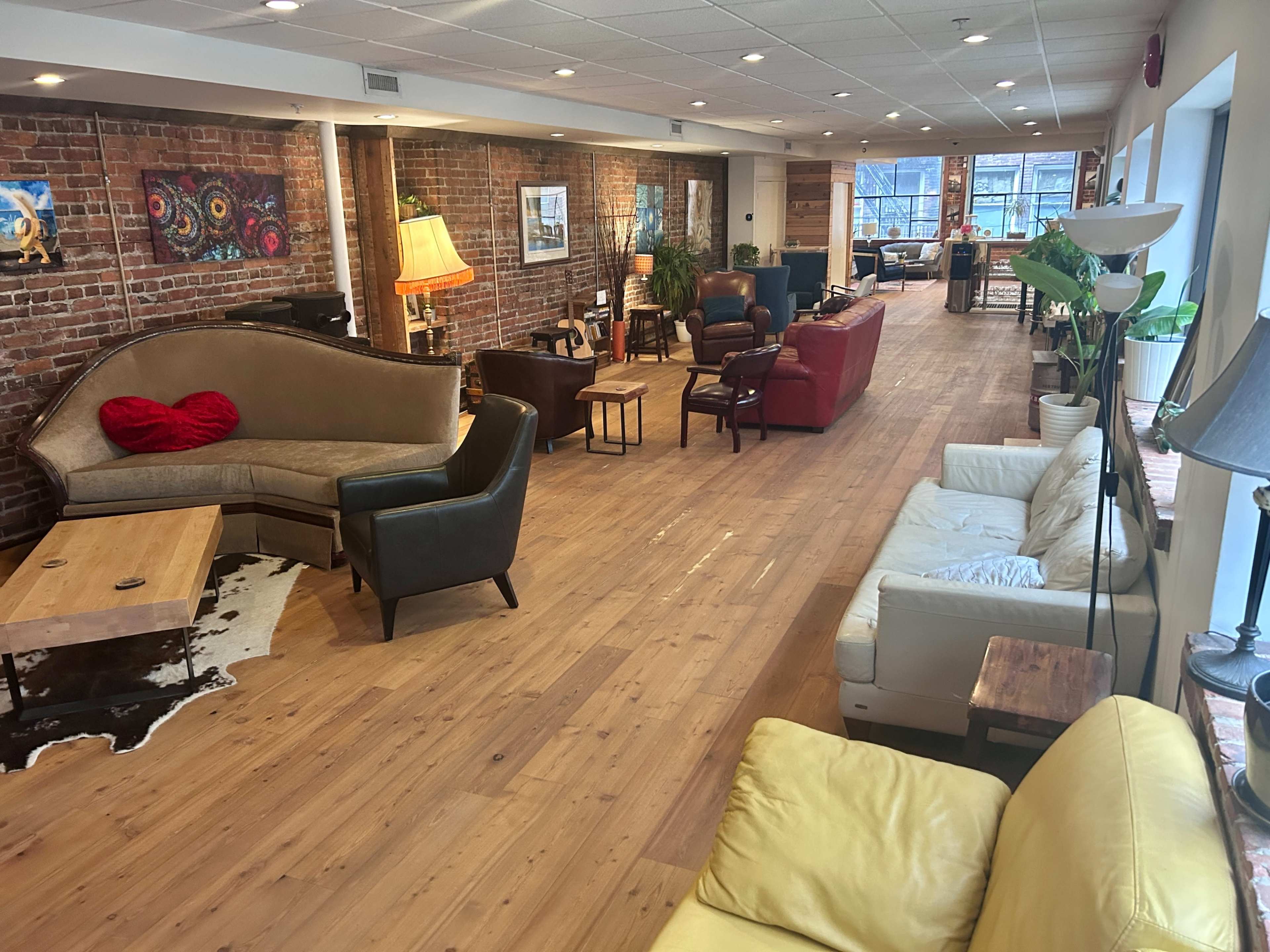 The image shows a spacious lounge area with various seating options, including sofas and armchairs, arranged along a hardwood floor with exposed brick walls.