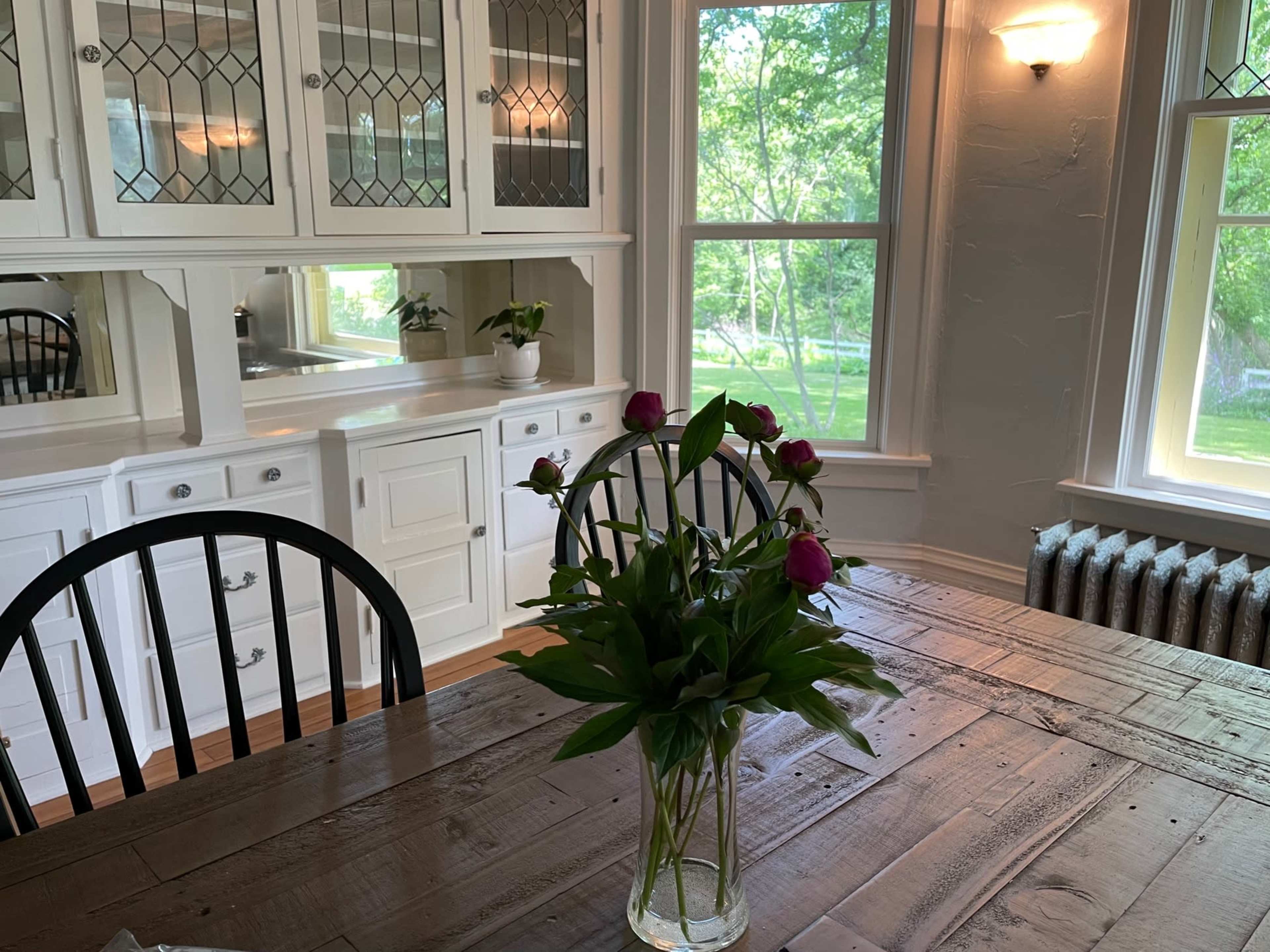 A dining area features a wooden table with a vase of flowers, surrounded by black chairs, and is illuminated by natural light from large windows.