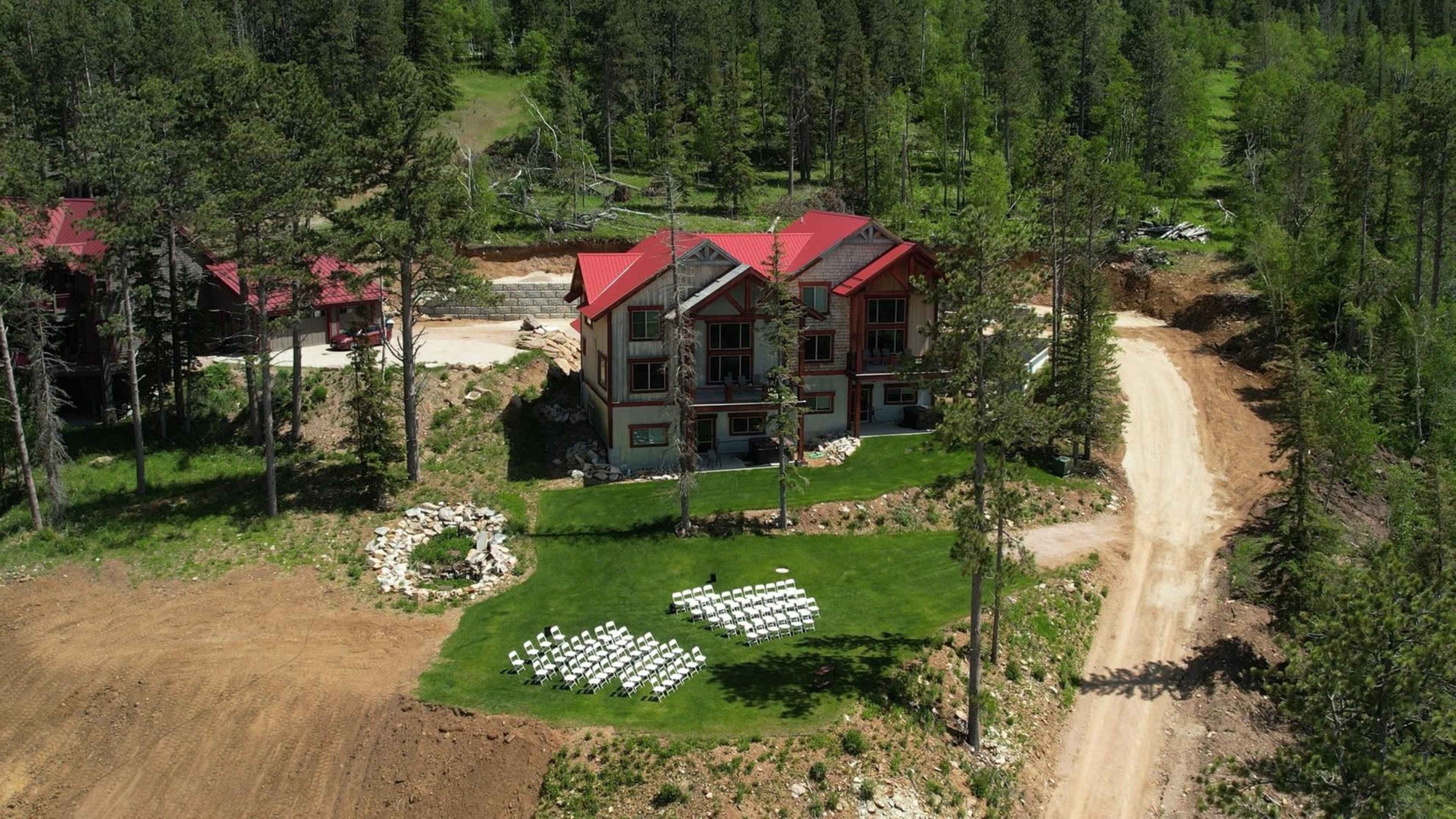 An aerial view shows a modern house surrounded by trees with a path leading away and a white seating arrangement set up on the lawn.