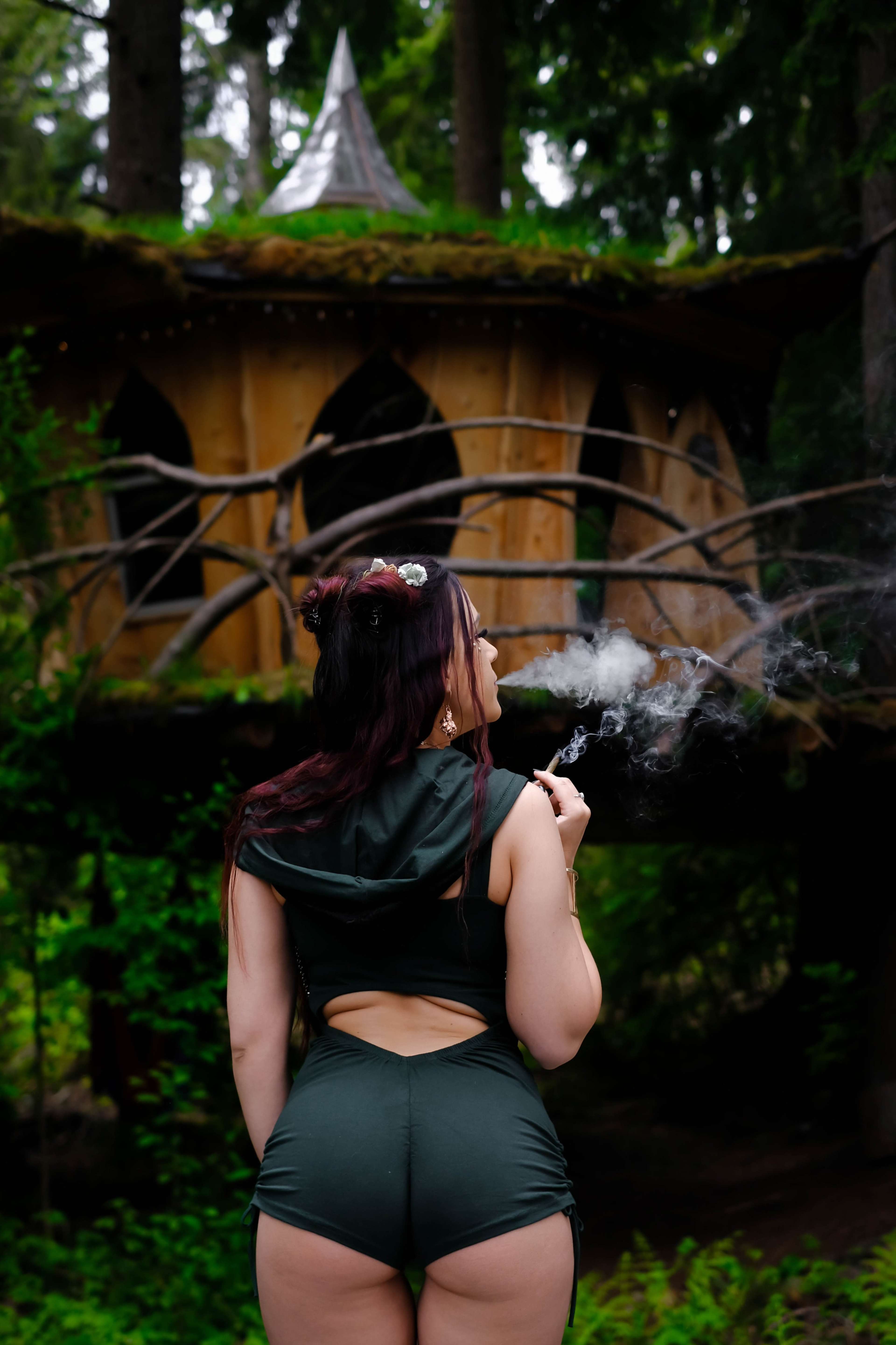 A person stands in front of a wooden structure surrounded by trees, smoking a cigarette.