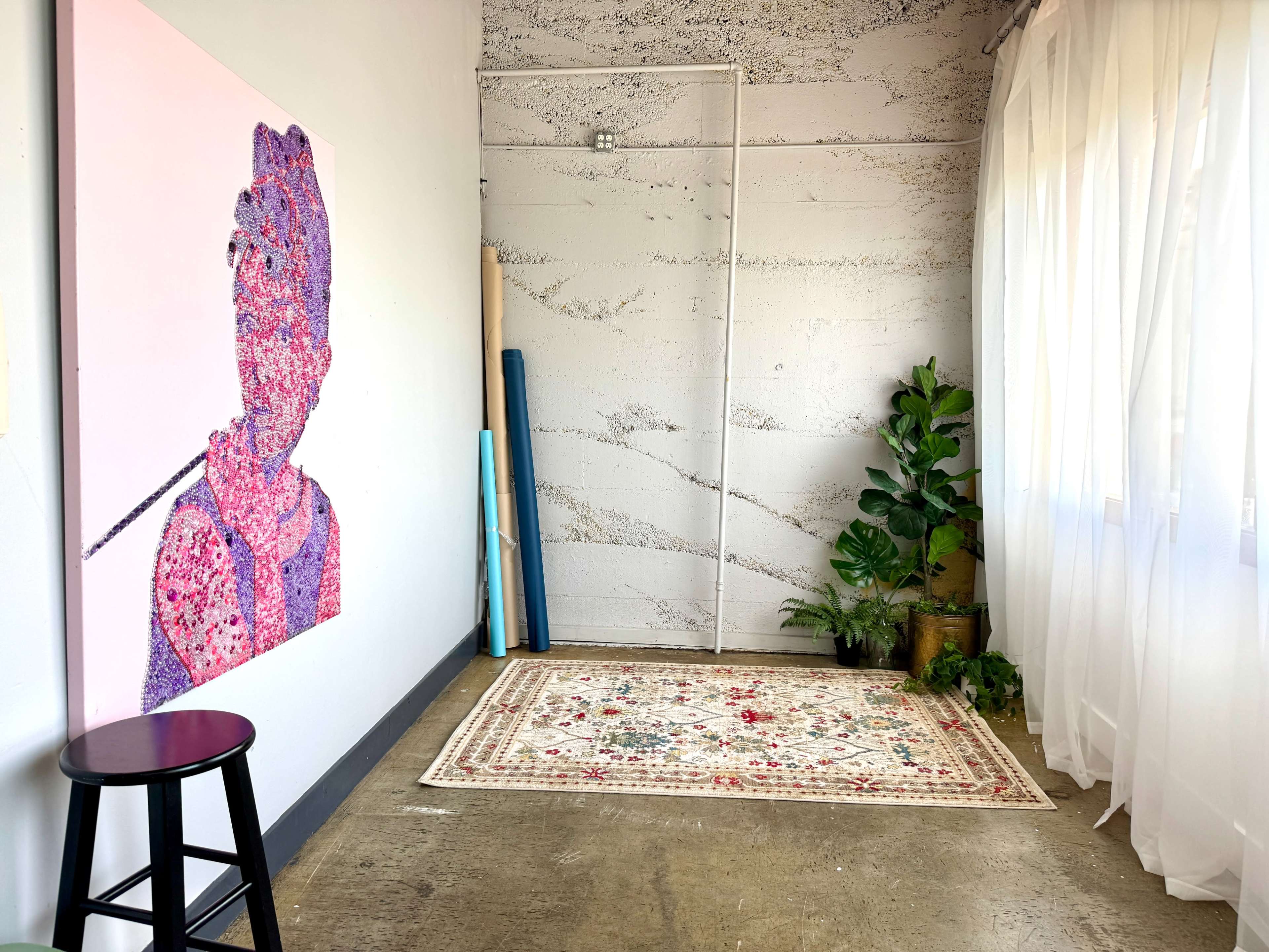 The image shows a bright interior space featuring a large pink and purple artwork on the wall, a decorative rug on the floor, a stool, and a potted plant near the window.