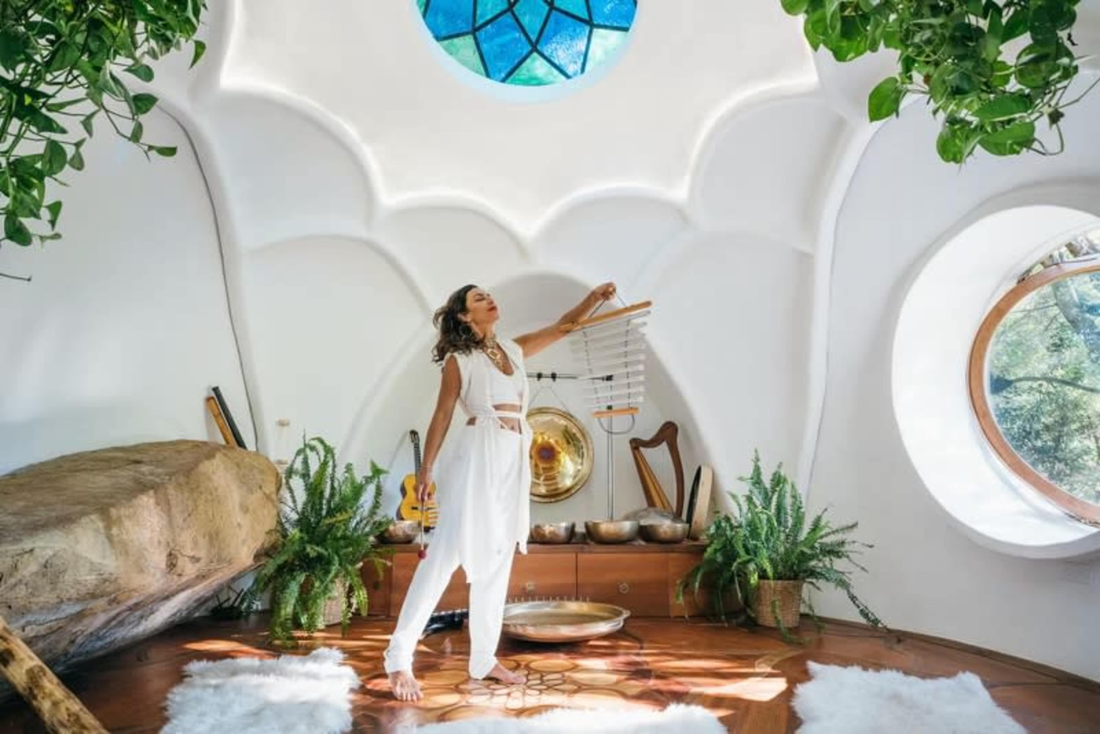 A woman in white clothing stands in a brightly lit, circular room with natural elements and musical instruments.