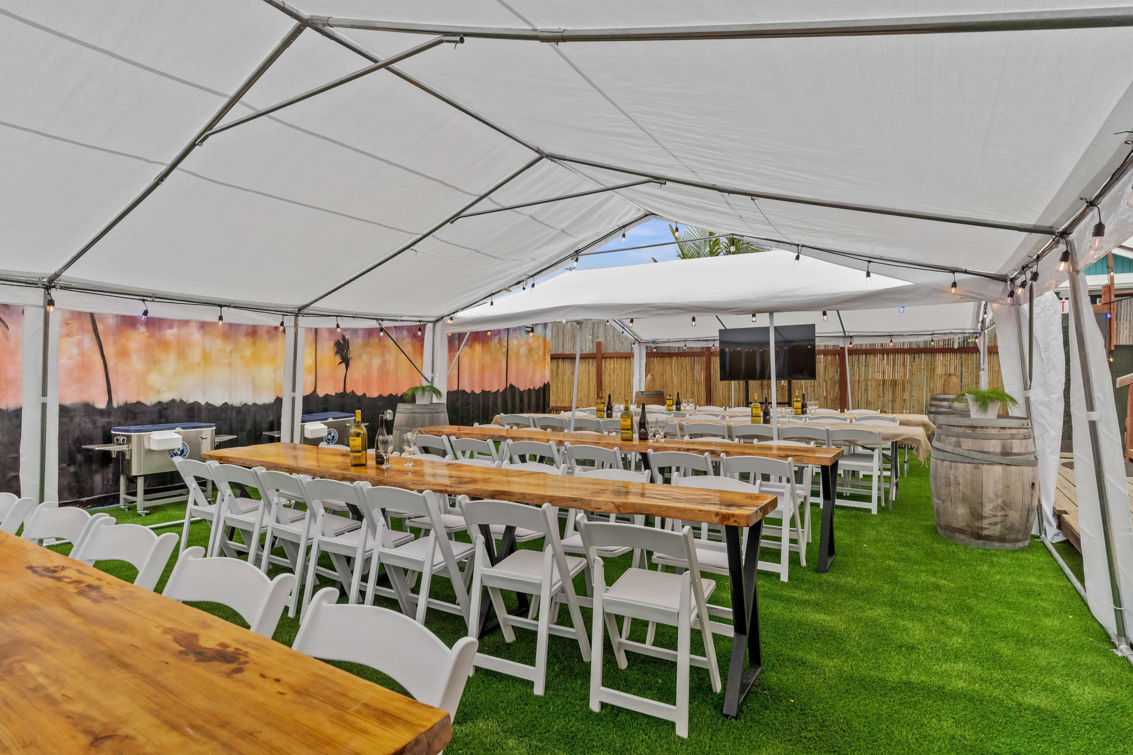 The image shows a large outdoor tent set up with wooden tables and white chairs arranged on green artificial grass.