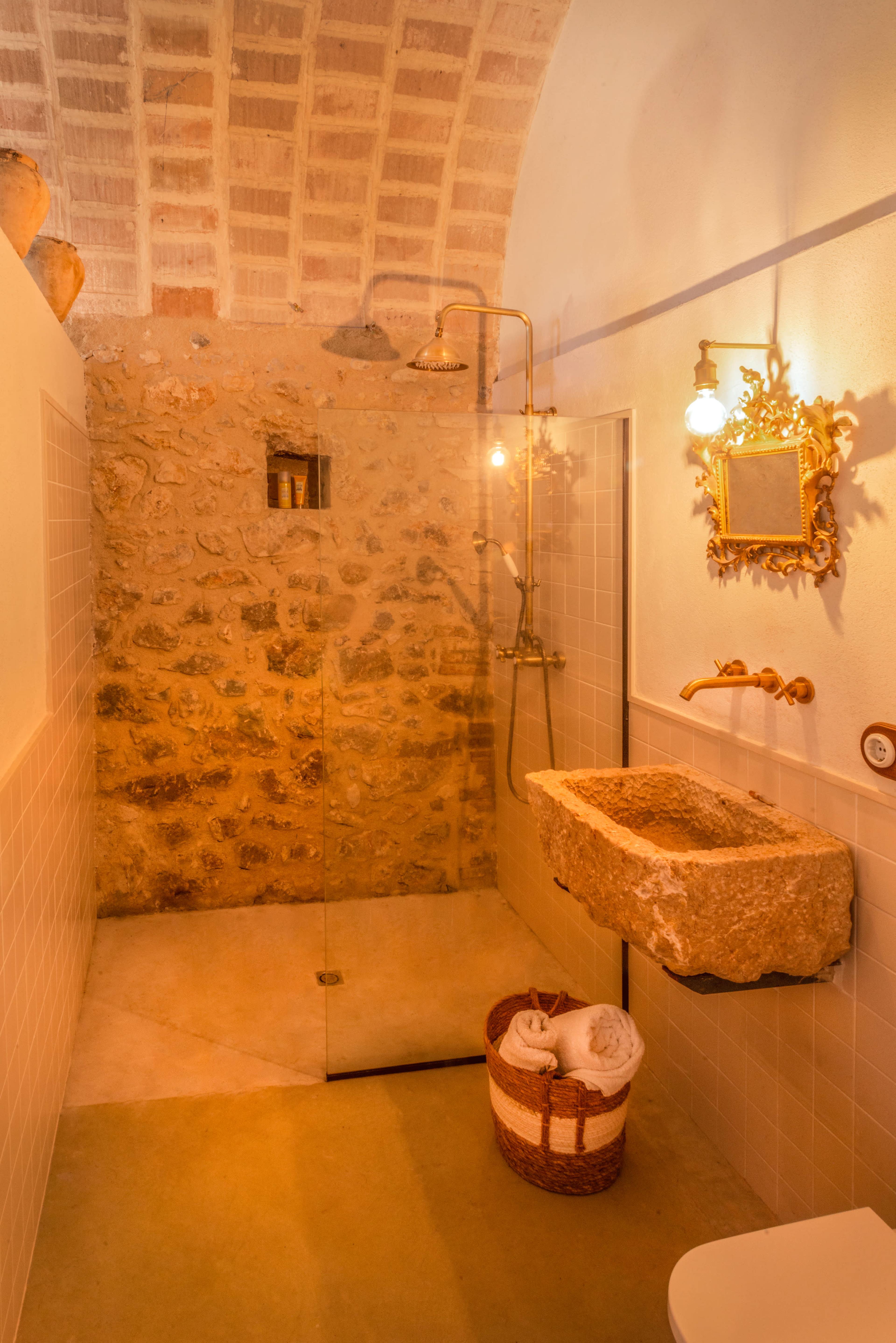 The image shows a bathroom featuring a stone sink, a glass shower enclosure, and a rustic stone wall, illuminated by warm lighting.