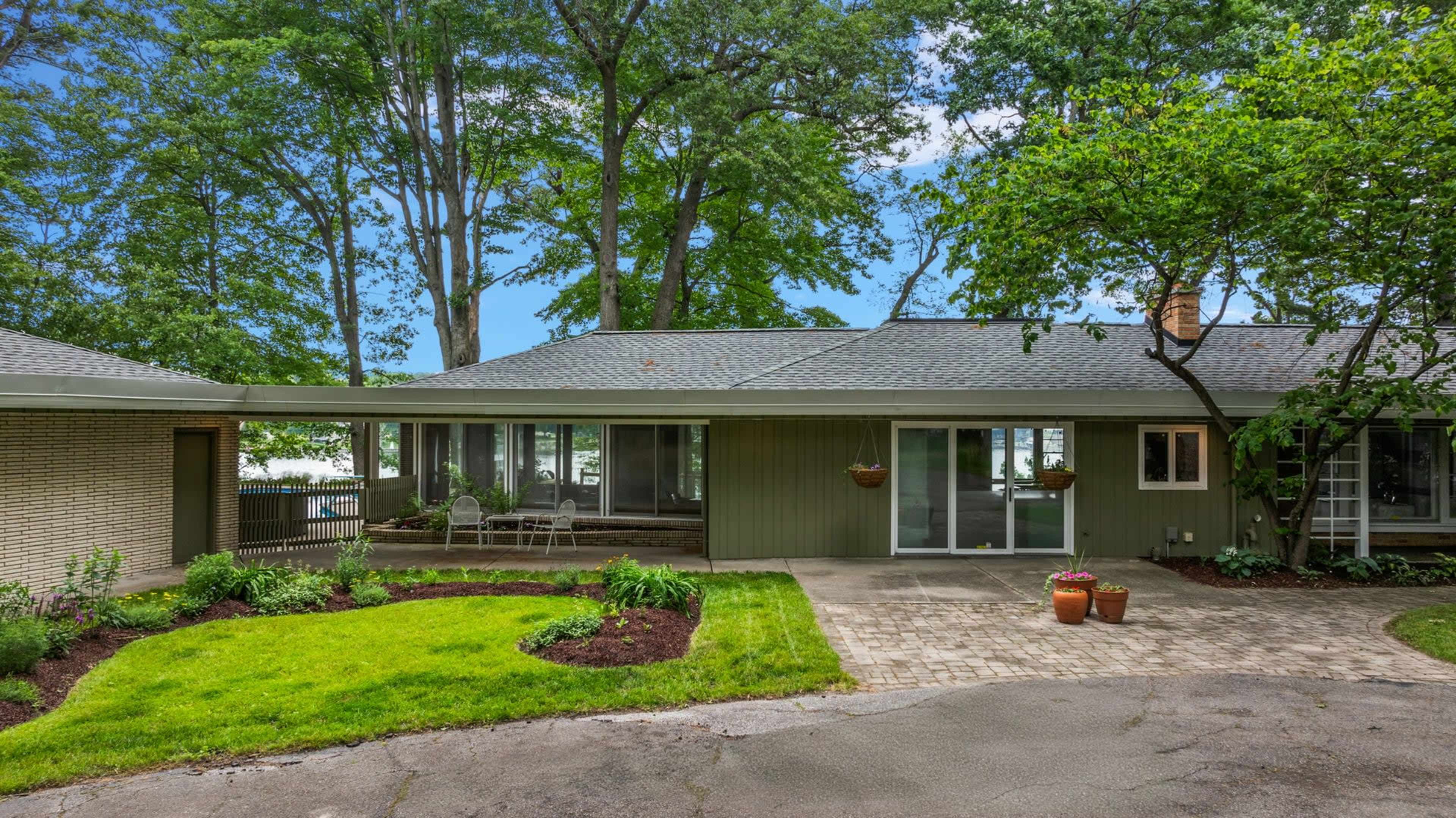 The image shows a single-story house with a green exterior, surrounded by trees and a landscaped yard featuring a paved driveway and flower pots.