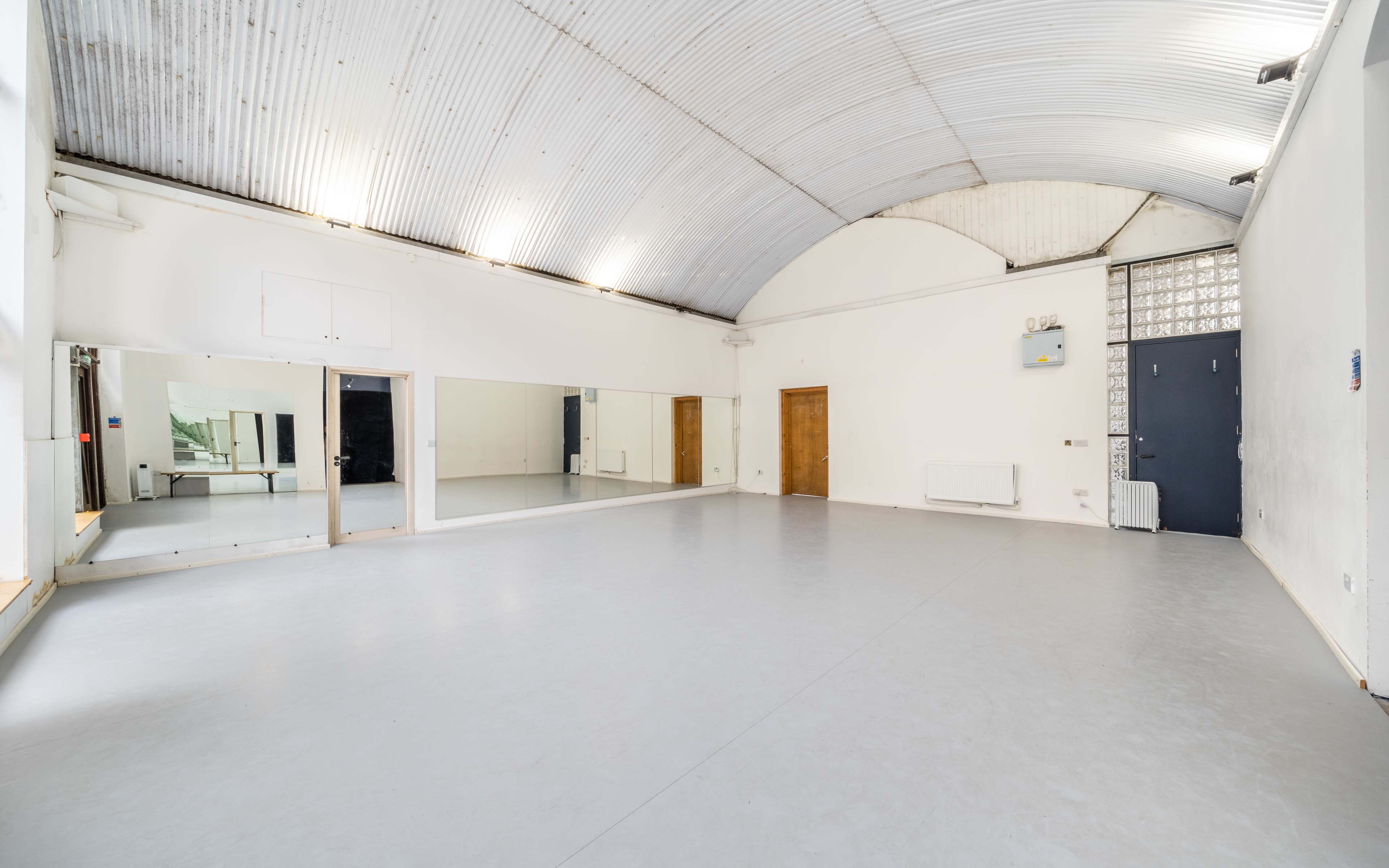 An empty studio space with a mirrored wall, wooden doors, and a high arched ceiling.