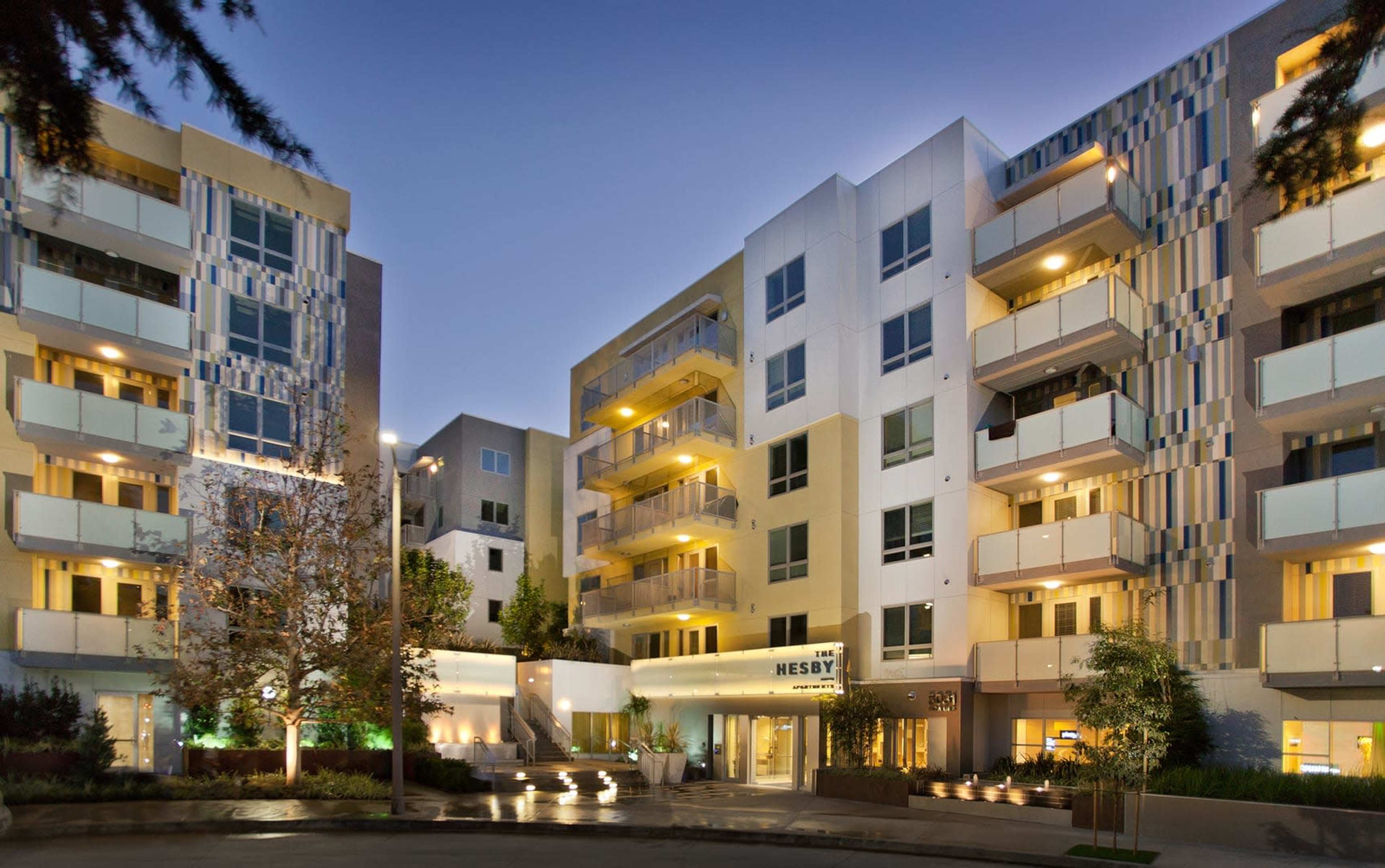 A modern apartment complex with multiple buildings illuminated at dusk, featuring balconies and landscaped areas.