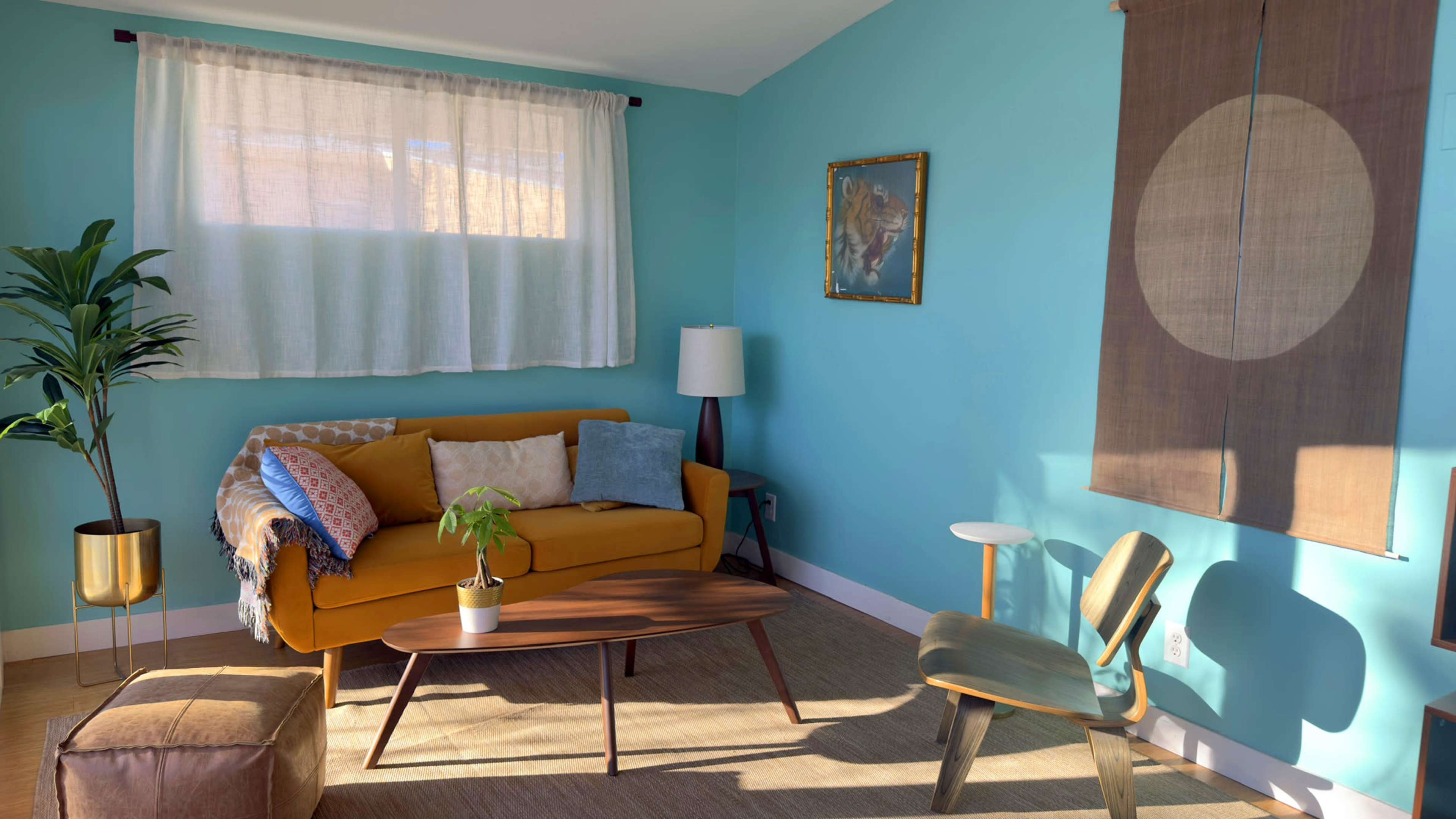 The room features a yellow sofa, a wooden coffee table, a modern chair, and a window with sheer curtains, all against a bright blue wall.