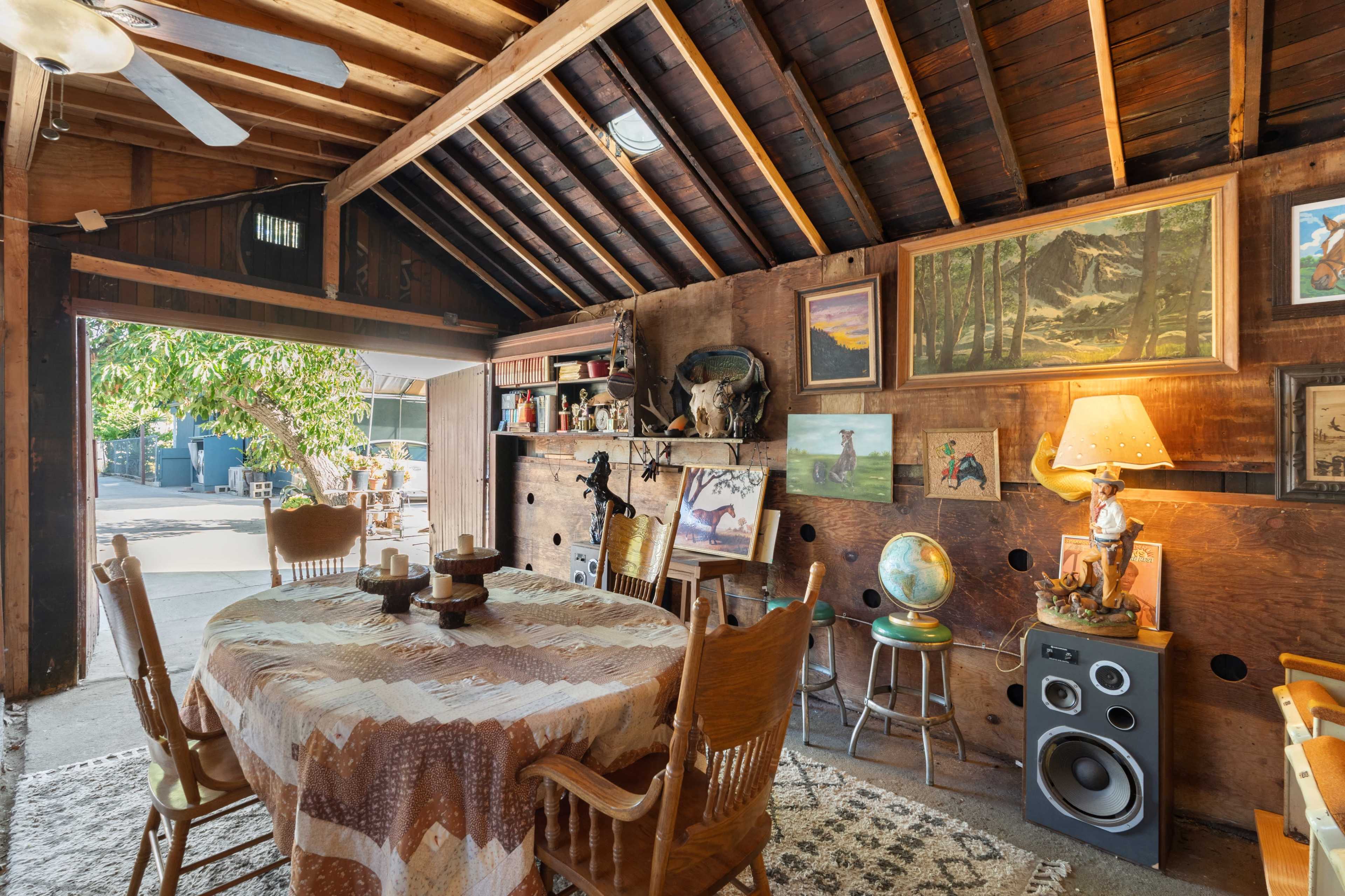 The image features a rustic interior of a barn-like space with a wooden ceiling, a round table surrounded by chairs, and various artworks displayed on the walls.