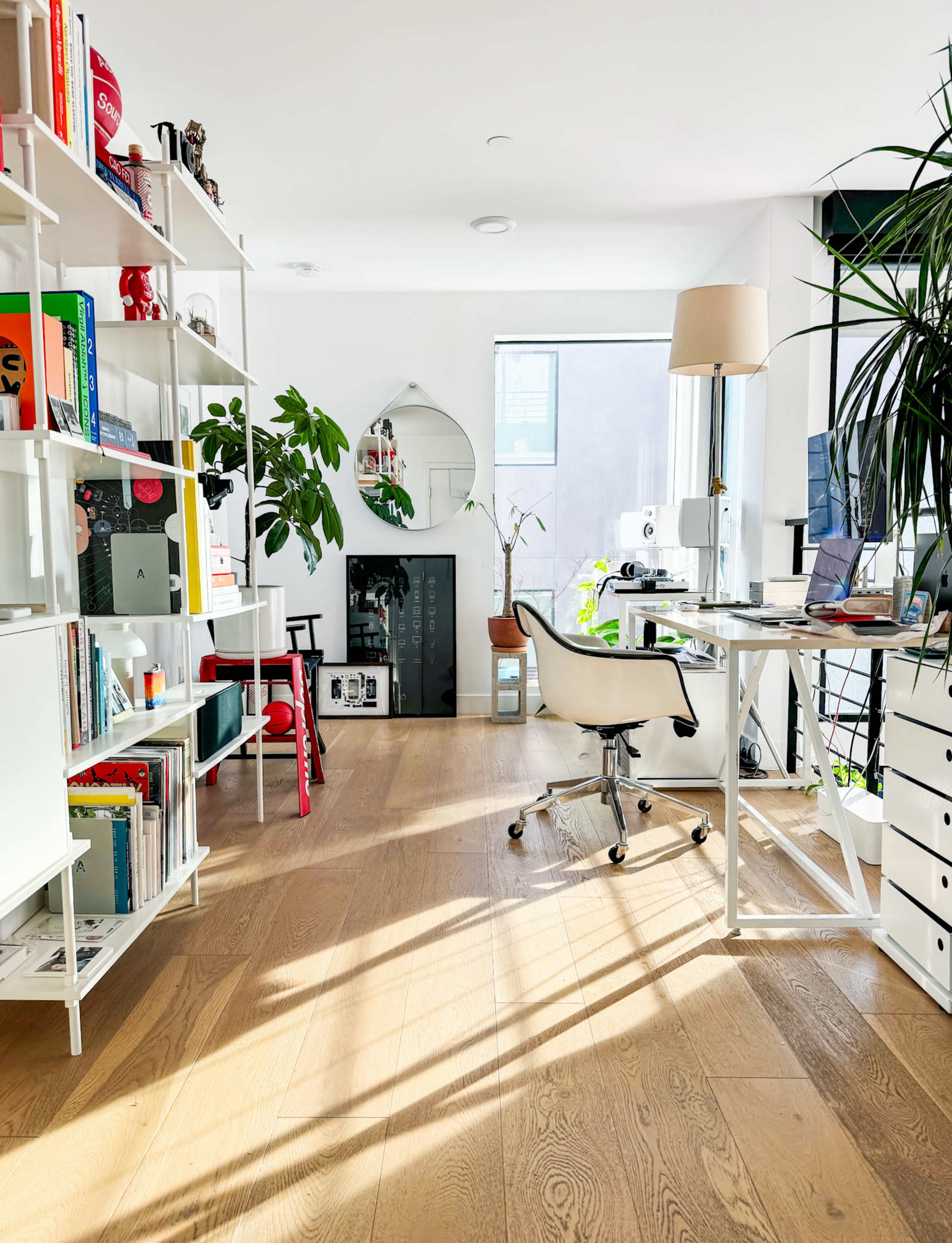 The image shows a bright modern office space with white bookshelves, a desk with a computer, and ample greenery.