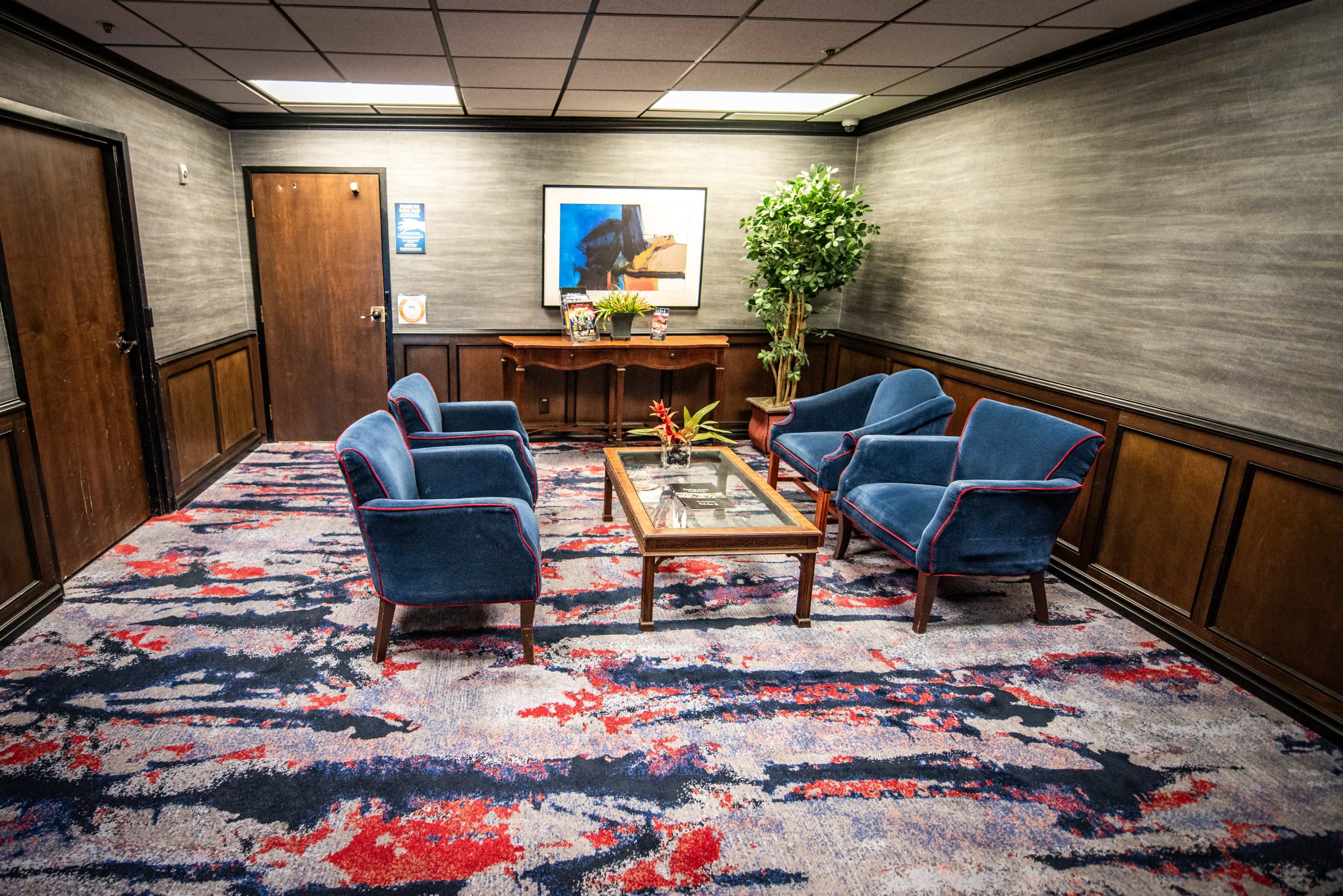 The image shows a waiting area with blue armchairs, a glass-top coffee table, and a potted plant against a textured wall with a piece of art.