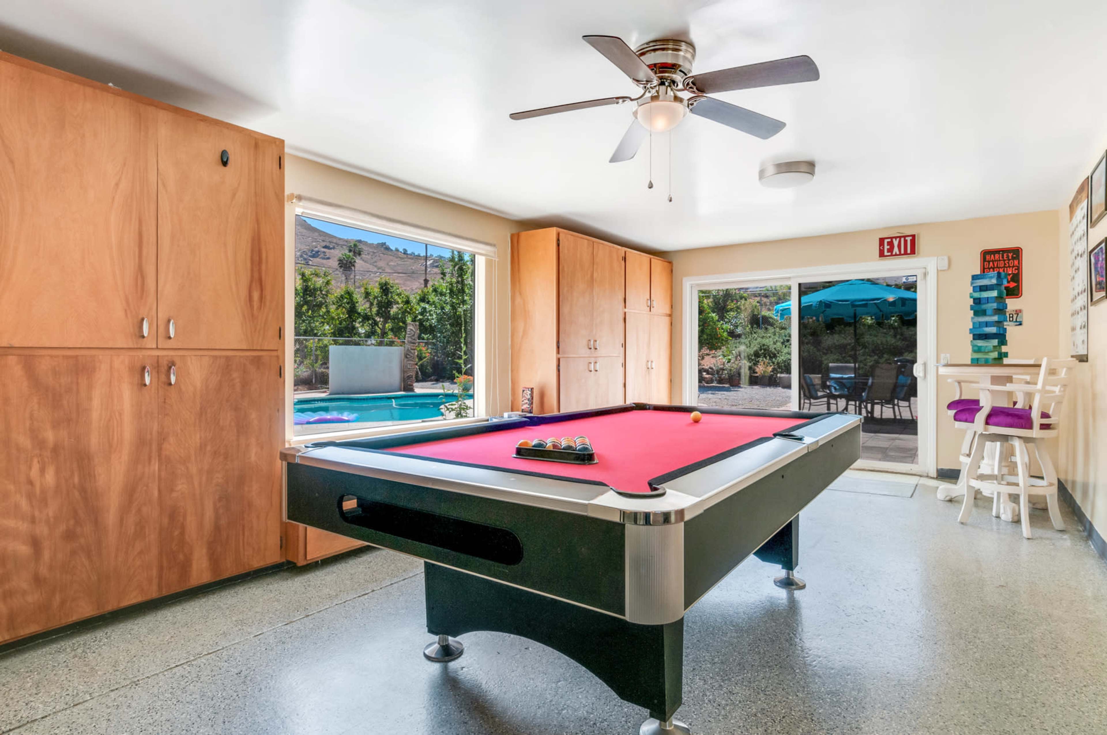 A red-felt pool table is positioned in a bright room with wooden cabinets and a view of a patio and pool area through glass doors.