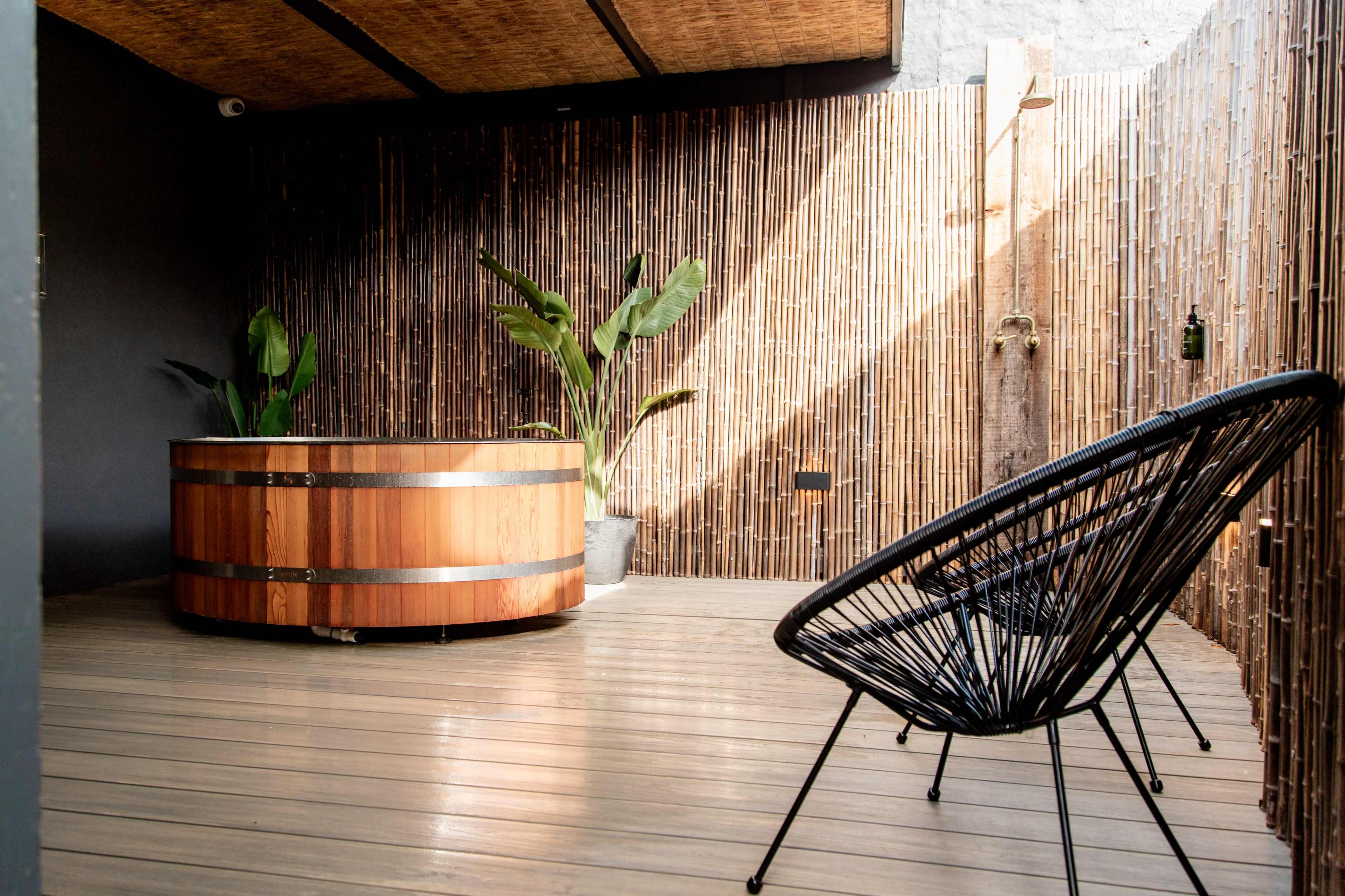 The image shows a wooden hot tub surrounded by bamboo walls and a black wire chair in a well-lit indoor space.