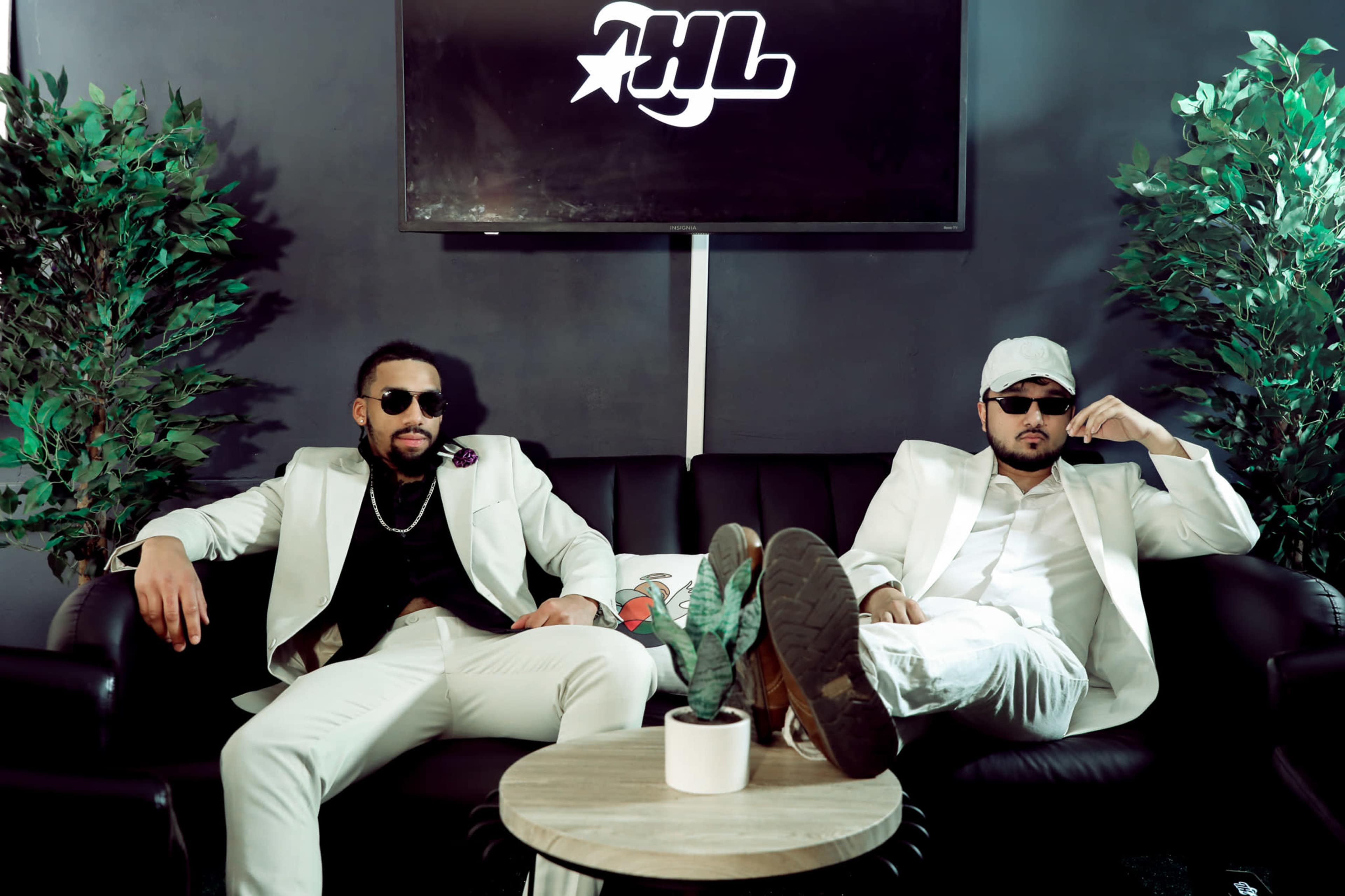 Two men in white suits sit on a black couch, with one wearing sunglasses and the other a cap, against a backdrop of green plants and a branded display.