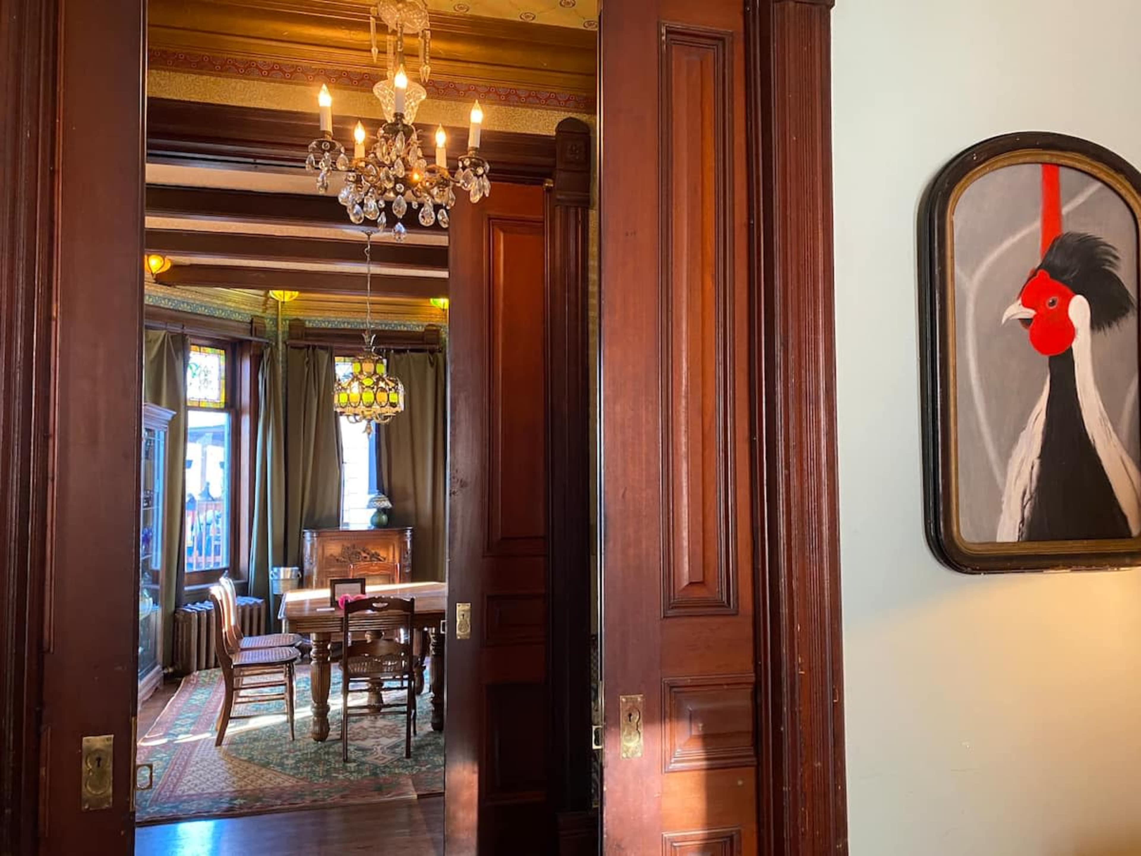 A grand wooden doorway reveals a well-furnished room featuring a chandelier, vintage furniture, and a wall-hung portrait of a rooster.