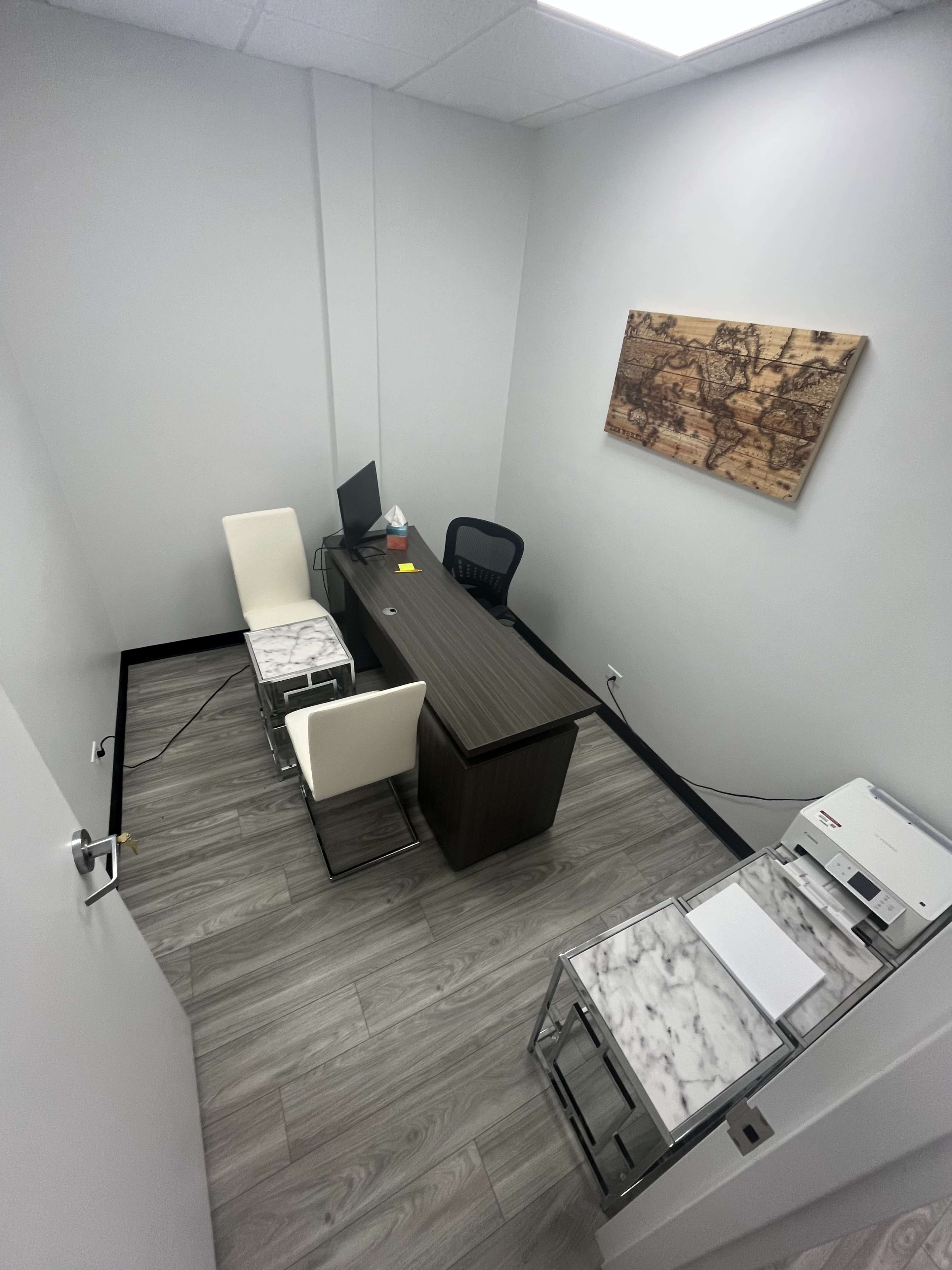 The image shows a small office space featuring a desk with a computer, two chairs, a printer, and a wall-mounted map.