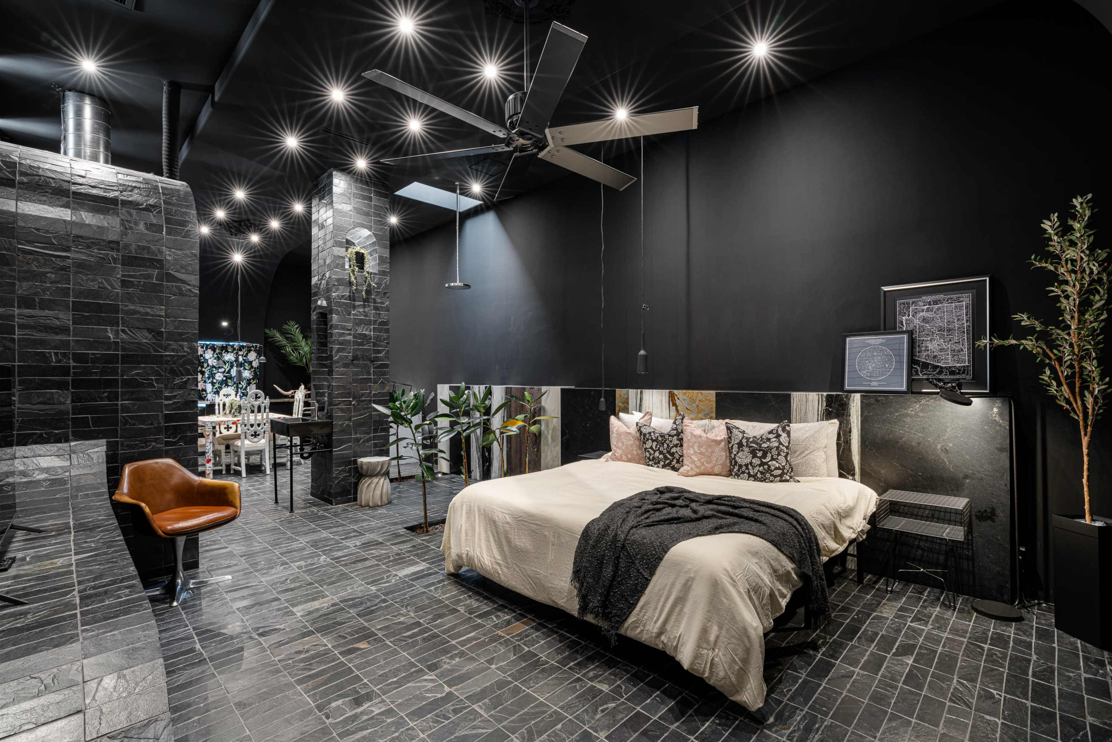 A modern bedroom with a large bed, dark walls, and sleek tiled flooring, featuring a ceiling fan and various indoor plants.