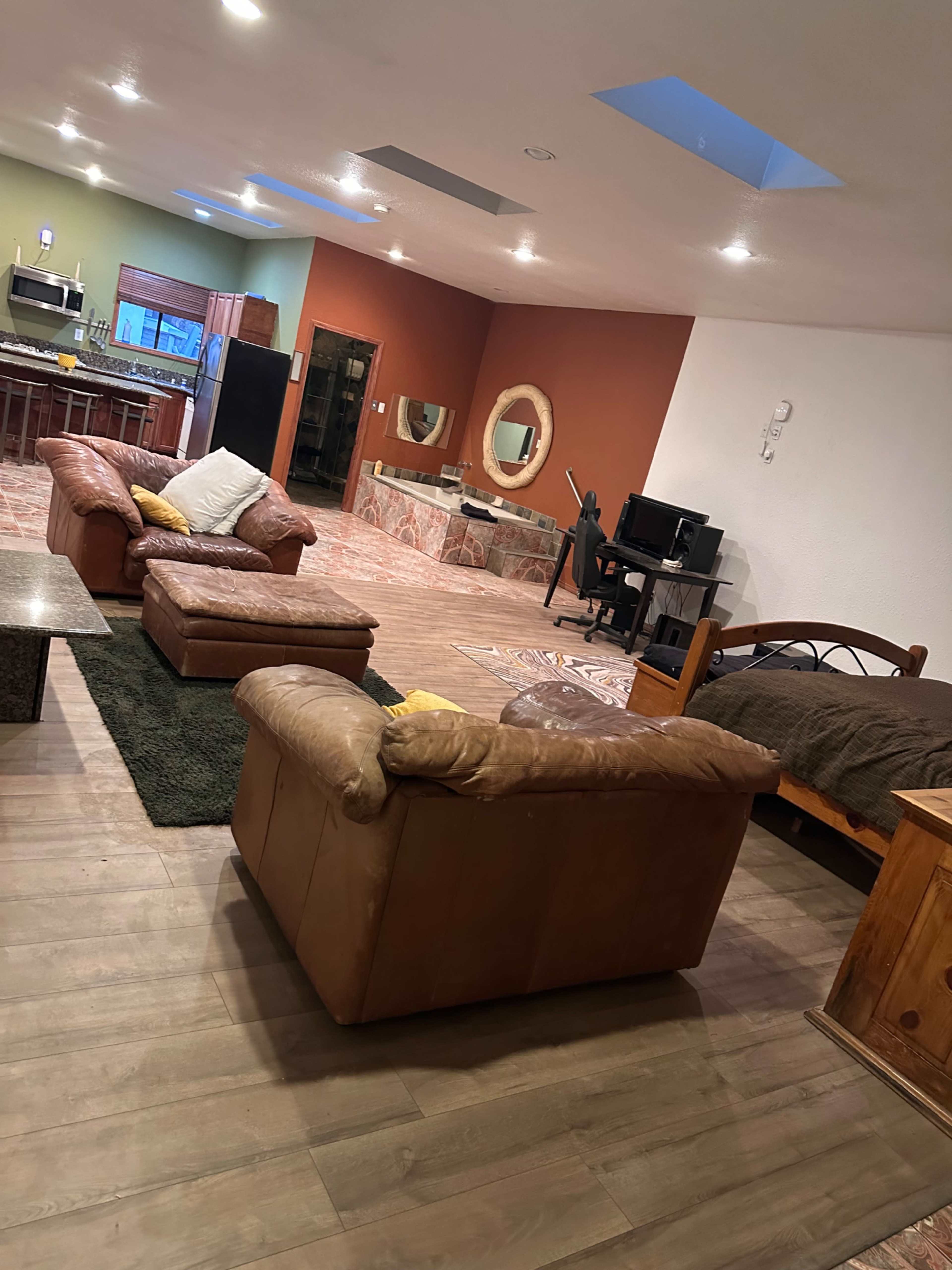 The image shows a spacious open living area featuring a kitchen, seating with two brown leather sofas, a bed, and a desk with a computer.