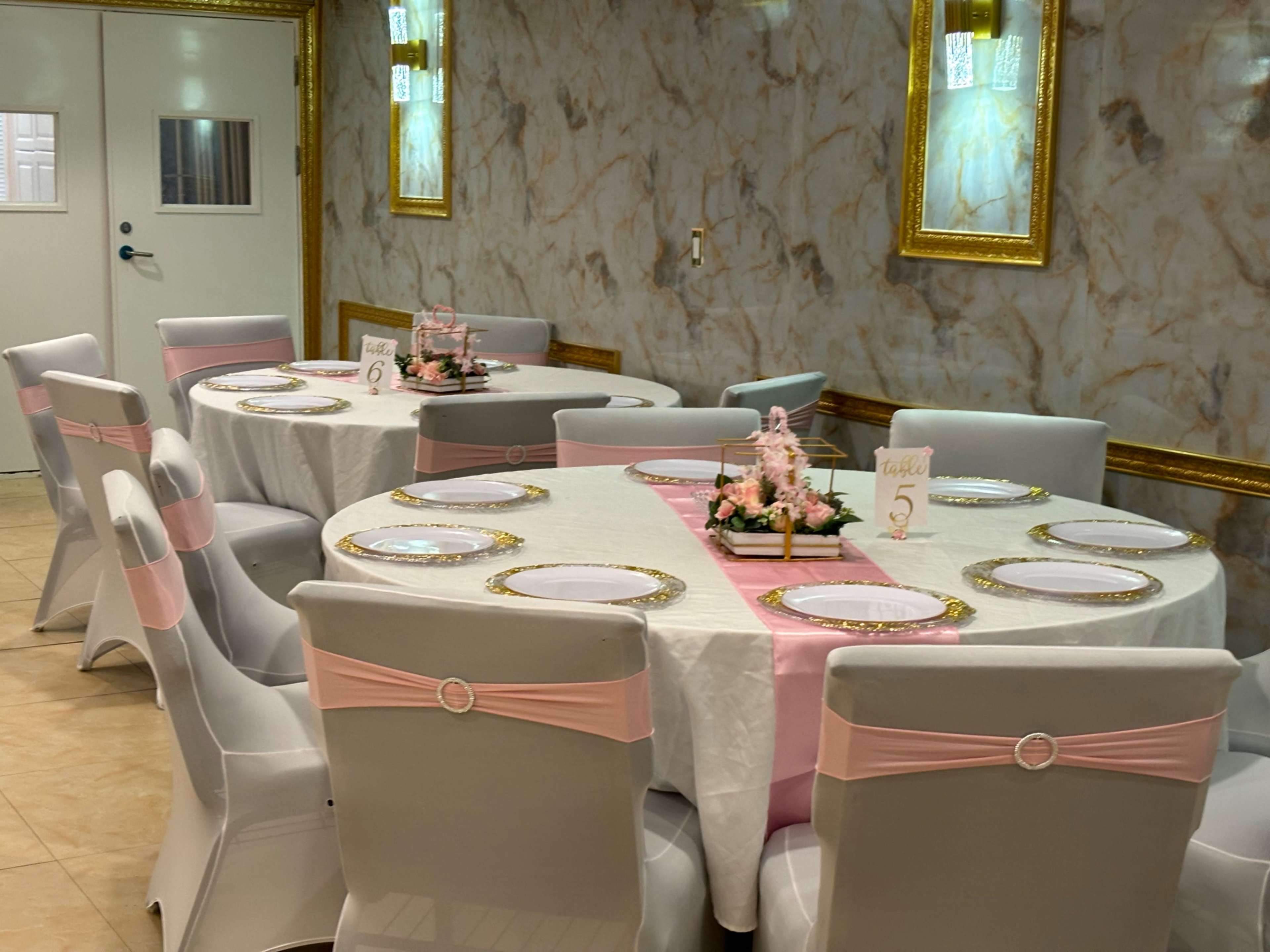 The image shows a banquet room featuring several circular tables covered with white tablecloths and decorated with pink accents, arranged in a spacious setting with marble walls and golden frames.