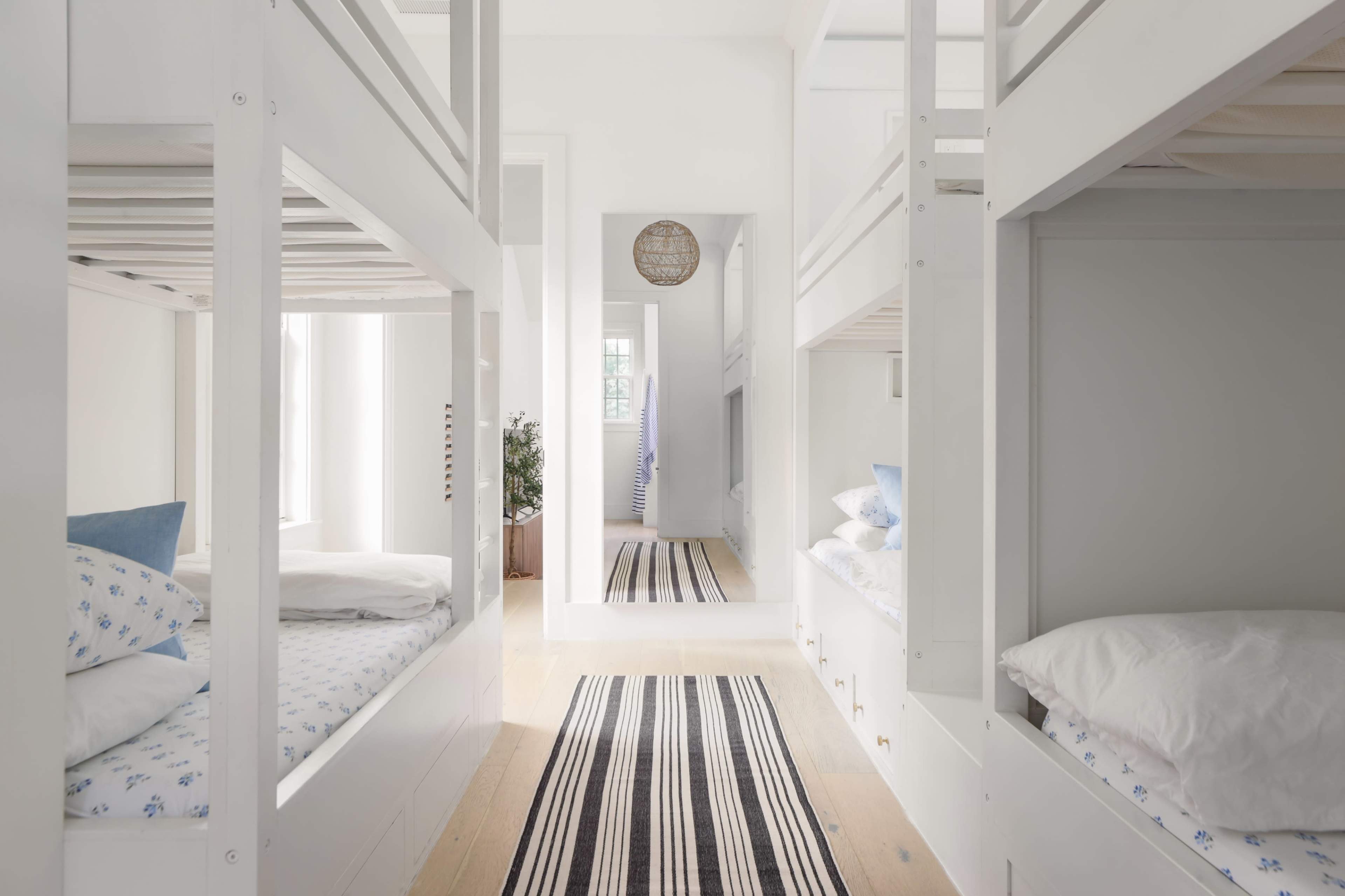 The image shows a bright and spacious room containing white bunk beds and a large mirror, with a striped rug running along the wooden floor.