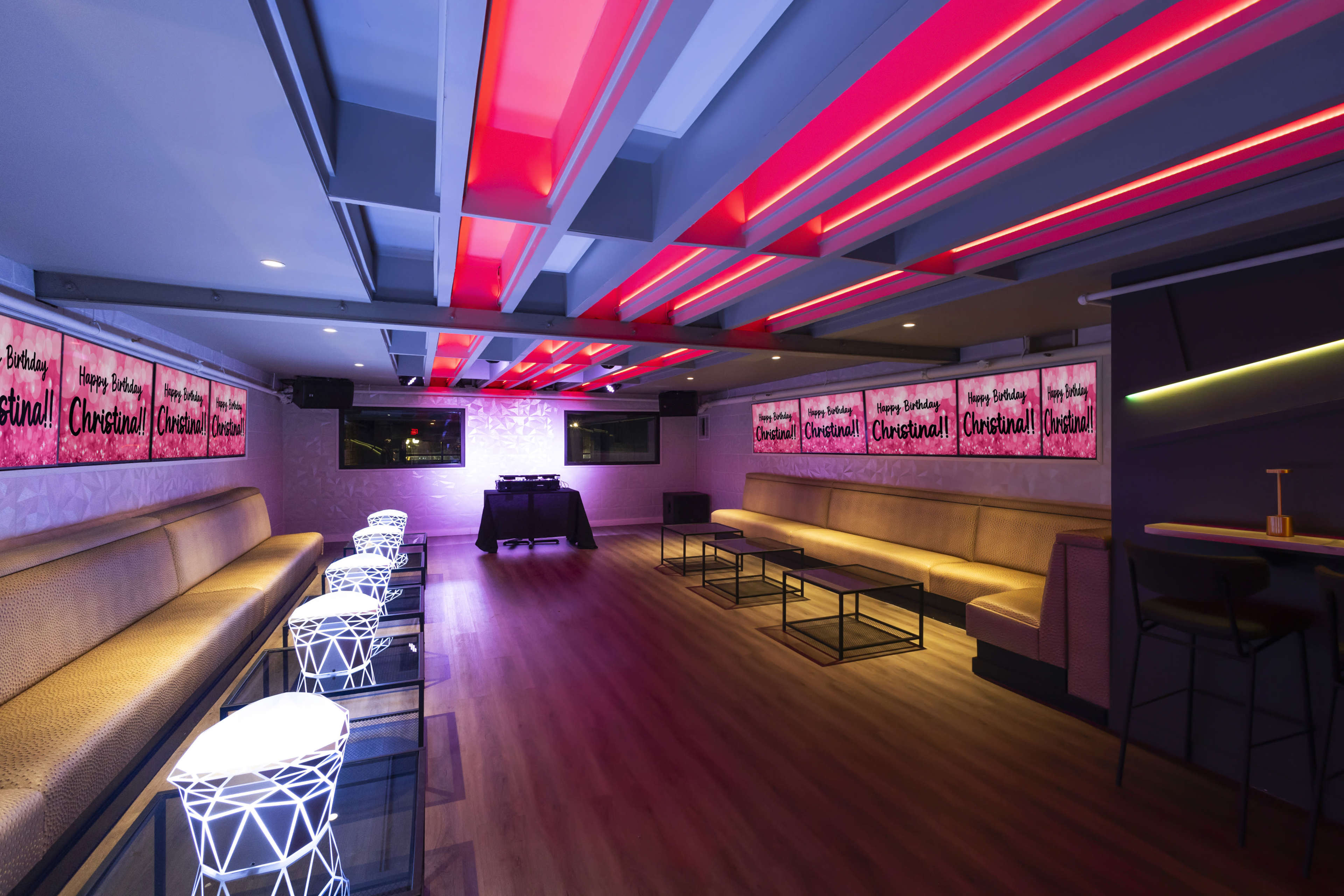 The image shows a modern lounge with illuminated seating, colorful light fixtures, and large screens displaying birthday messages.