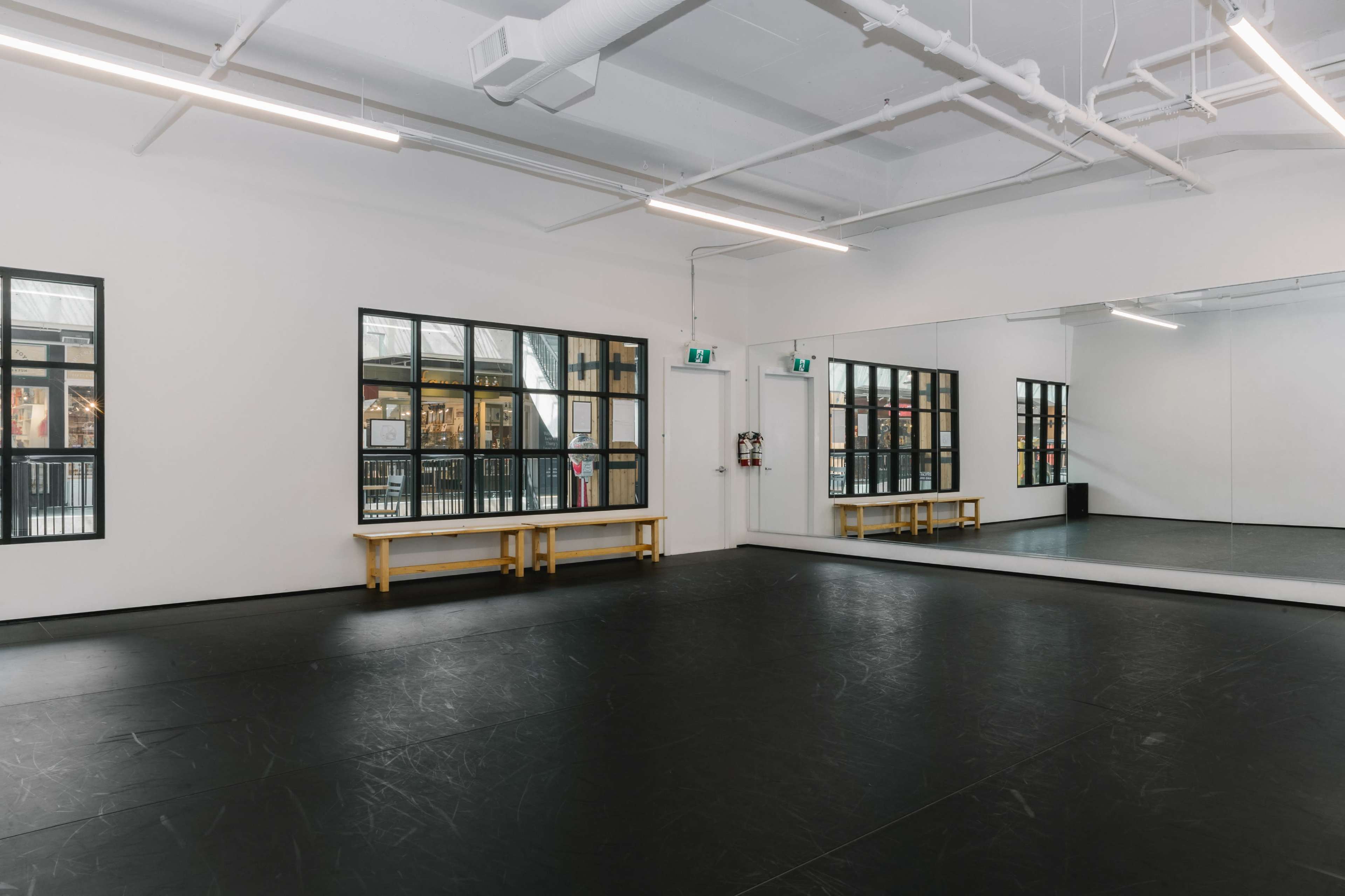 The image shows a spacious dance studio with black flooring, large windows allowing natural light, and a mirrored wall.