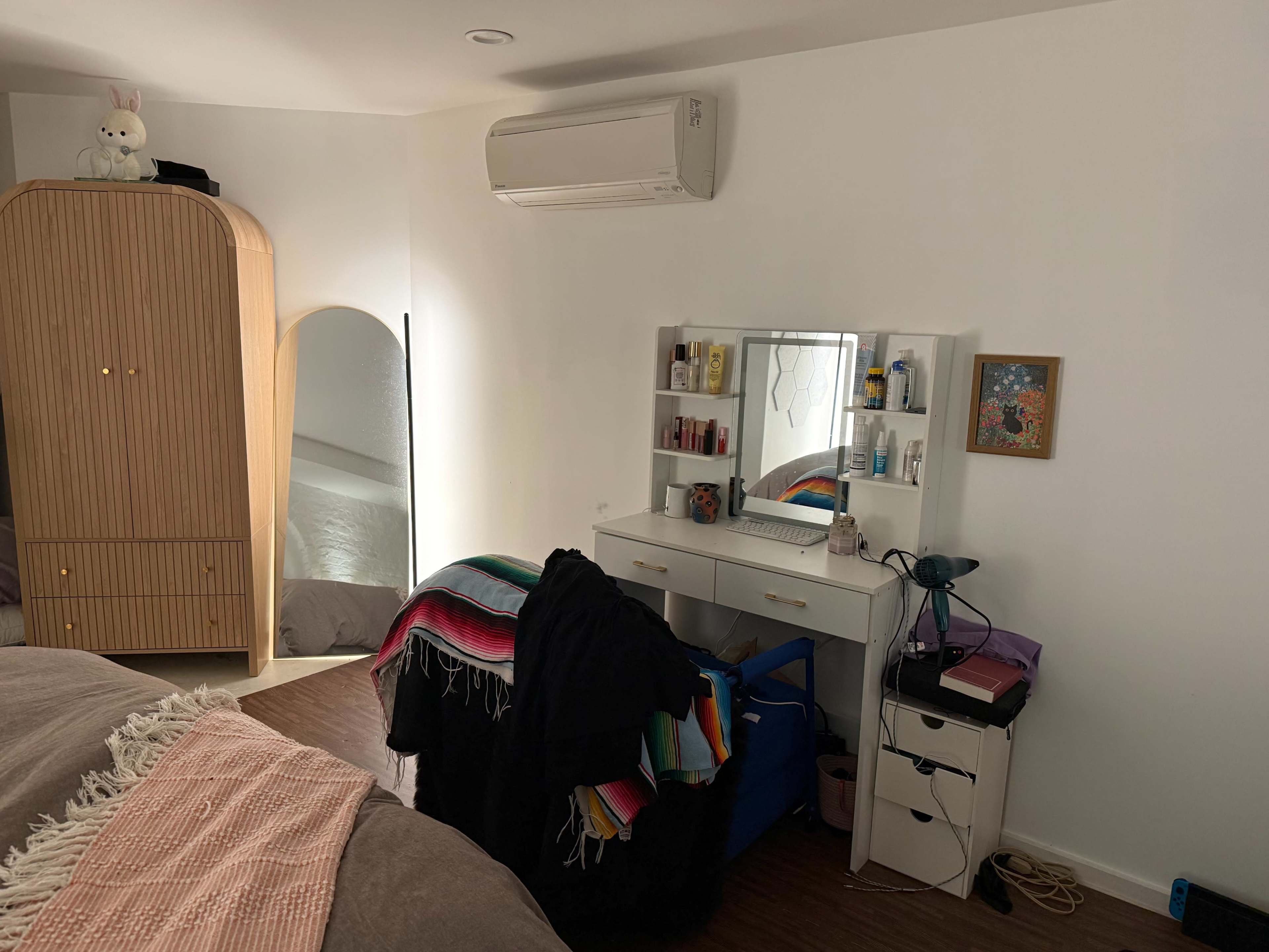 The image shows a bedroom with a bed in the foreground, a mirror and vanity area against the wall, and a closet with shelving in the background.