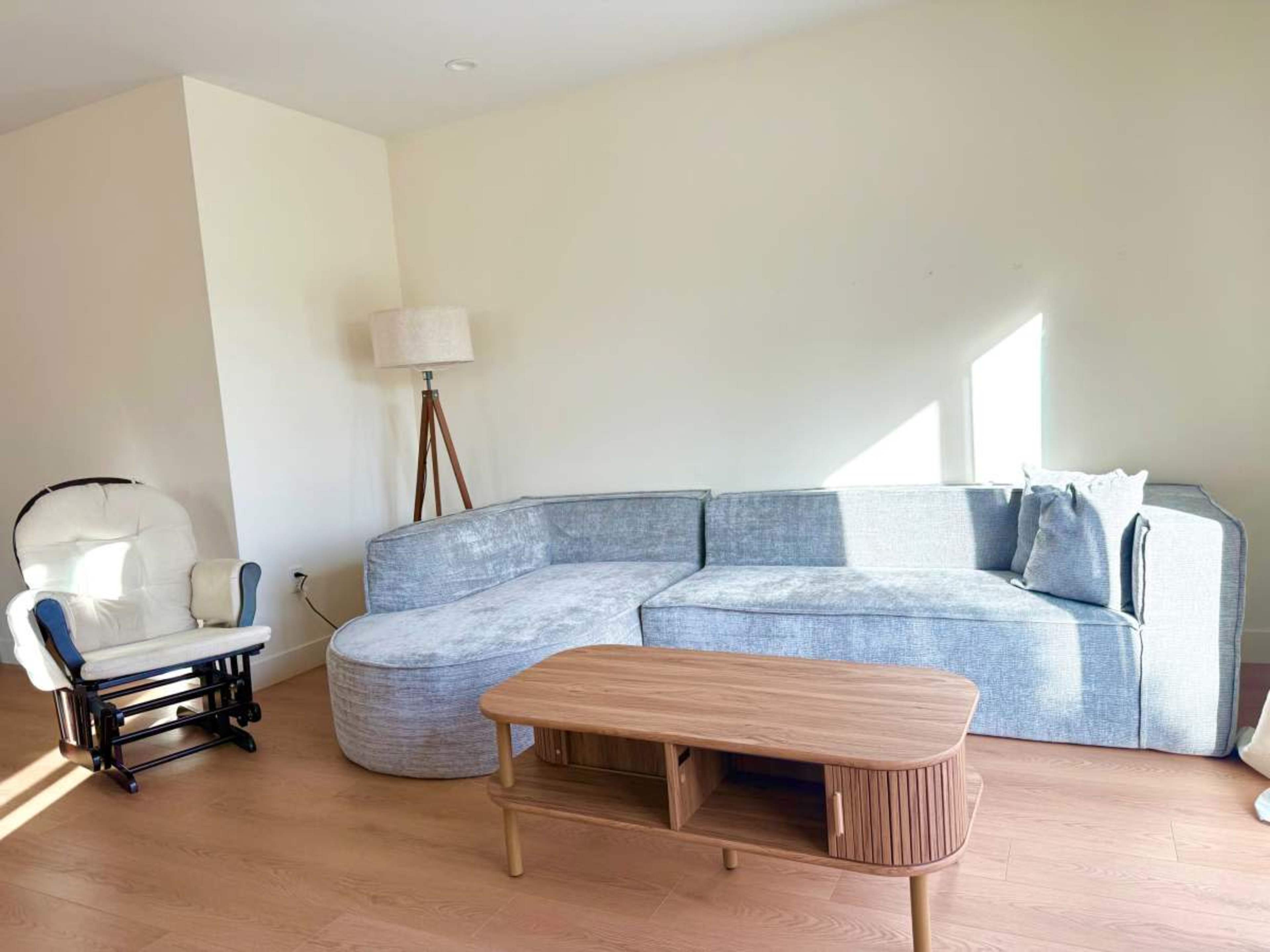 The image shows a modern living room with a light-colored sectional sofa, a wooden coffee table, and a rocking chair.