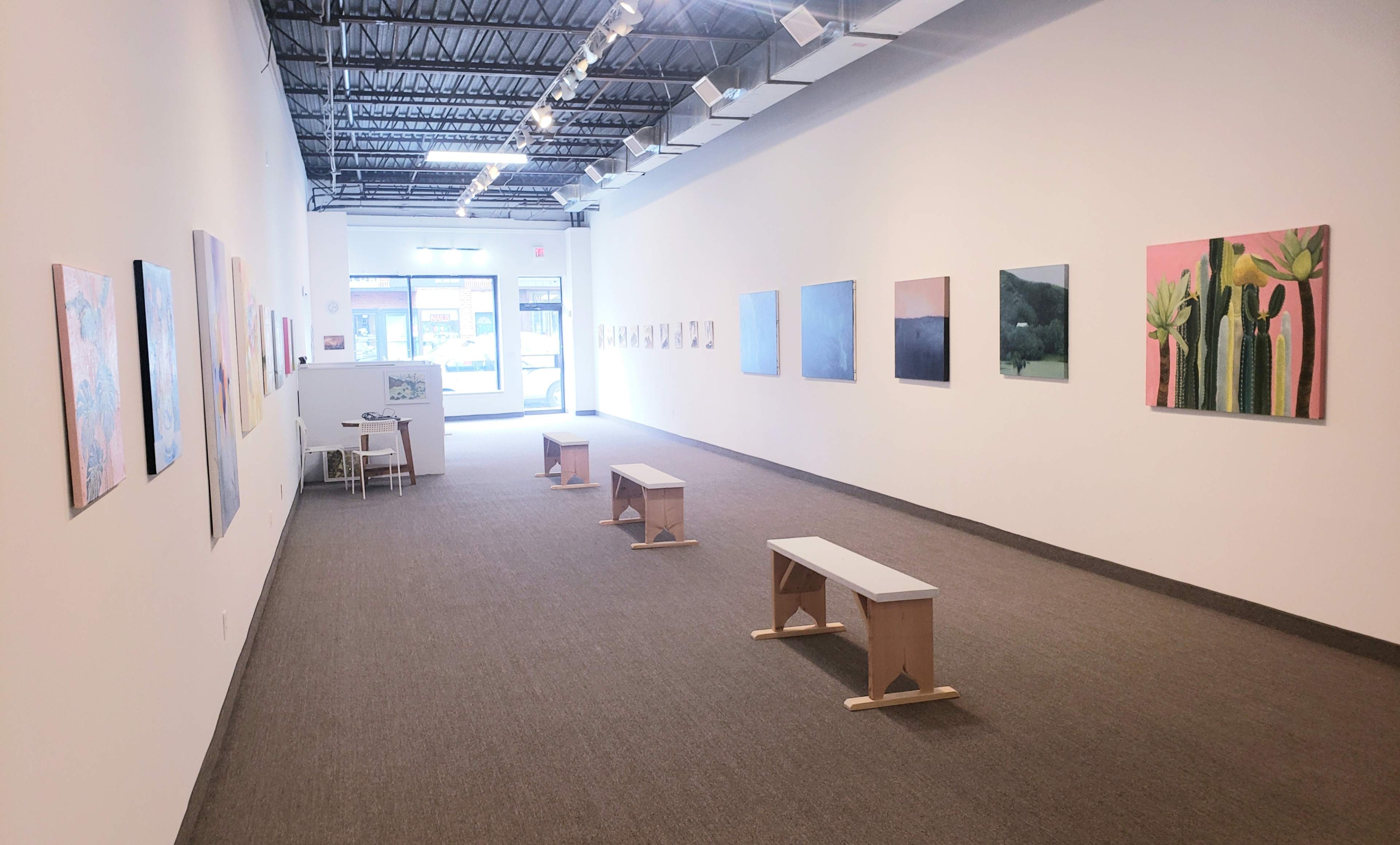 A spacious art gallery features white walls adorned with various paintings, with several benches arranged along the center and large windows illuminating the space.