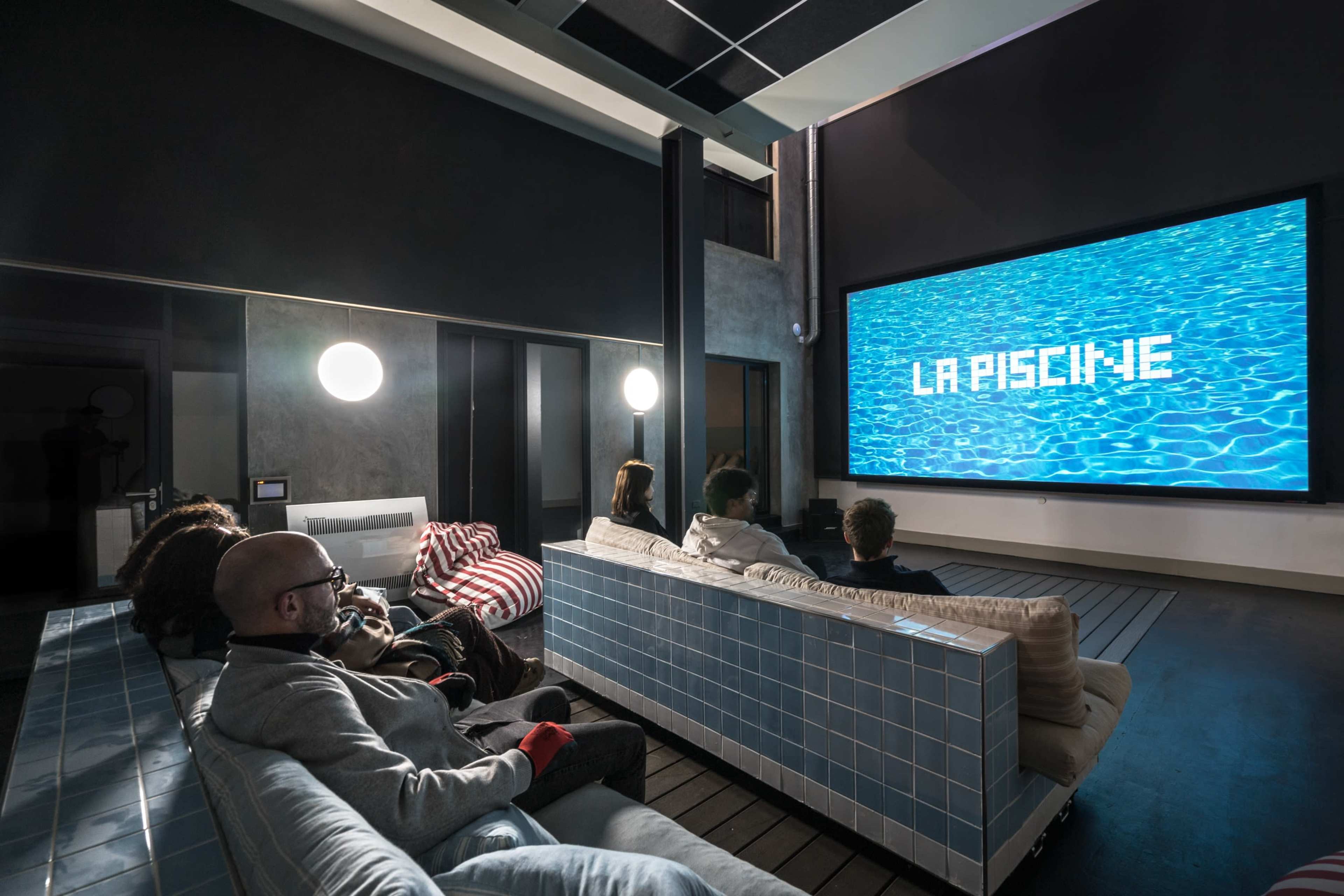 A group of six people sit on sofas in a modern home theater, watching a large screen displaying the words "LA PISCINE" over a blue water background.