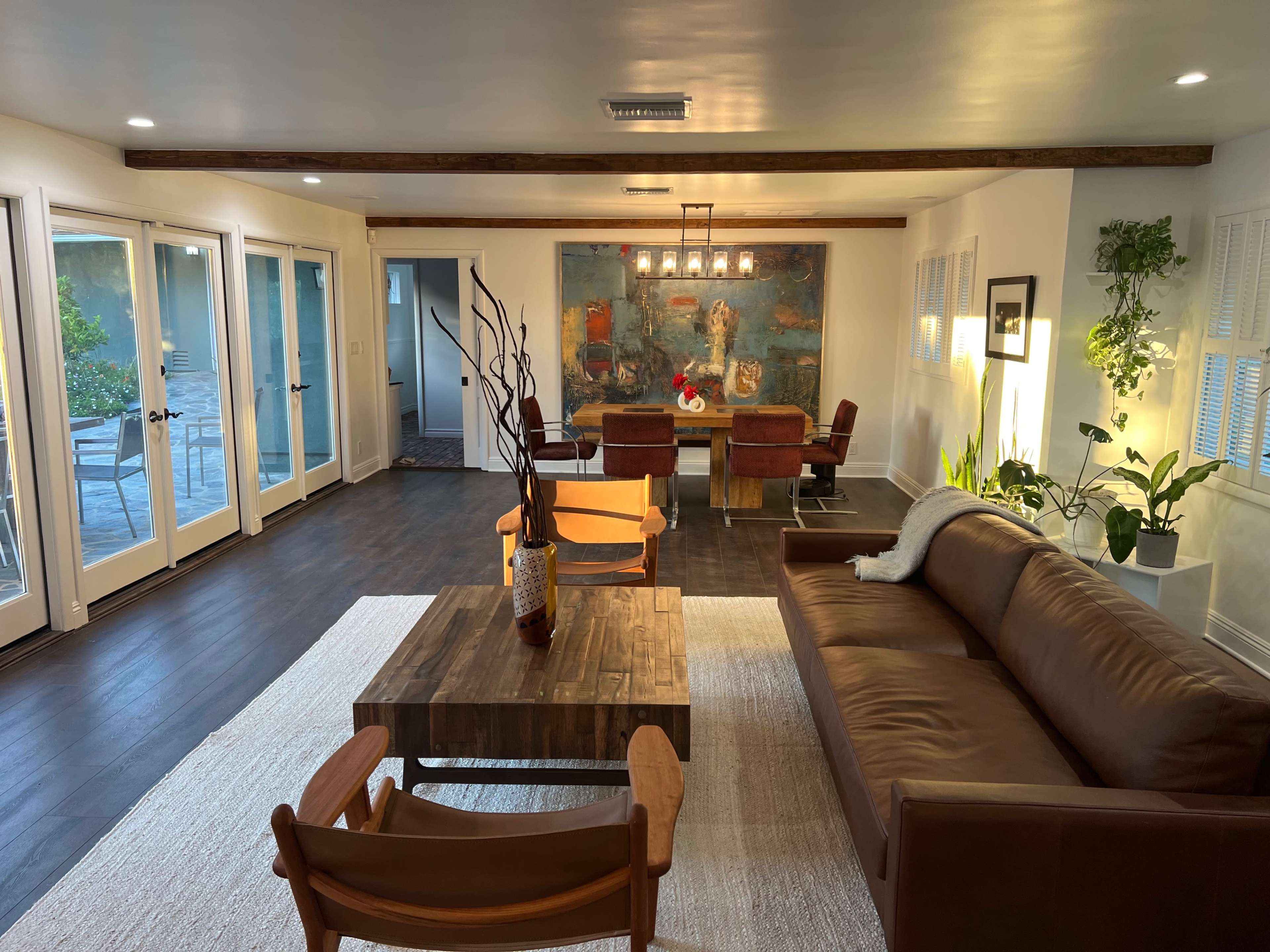 A cozy living room features a brown sofa, a wooden coffee table, and a pair of chairs, with large windows providing natural light and a view of a small patio.