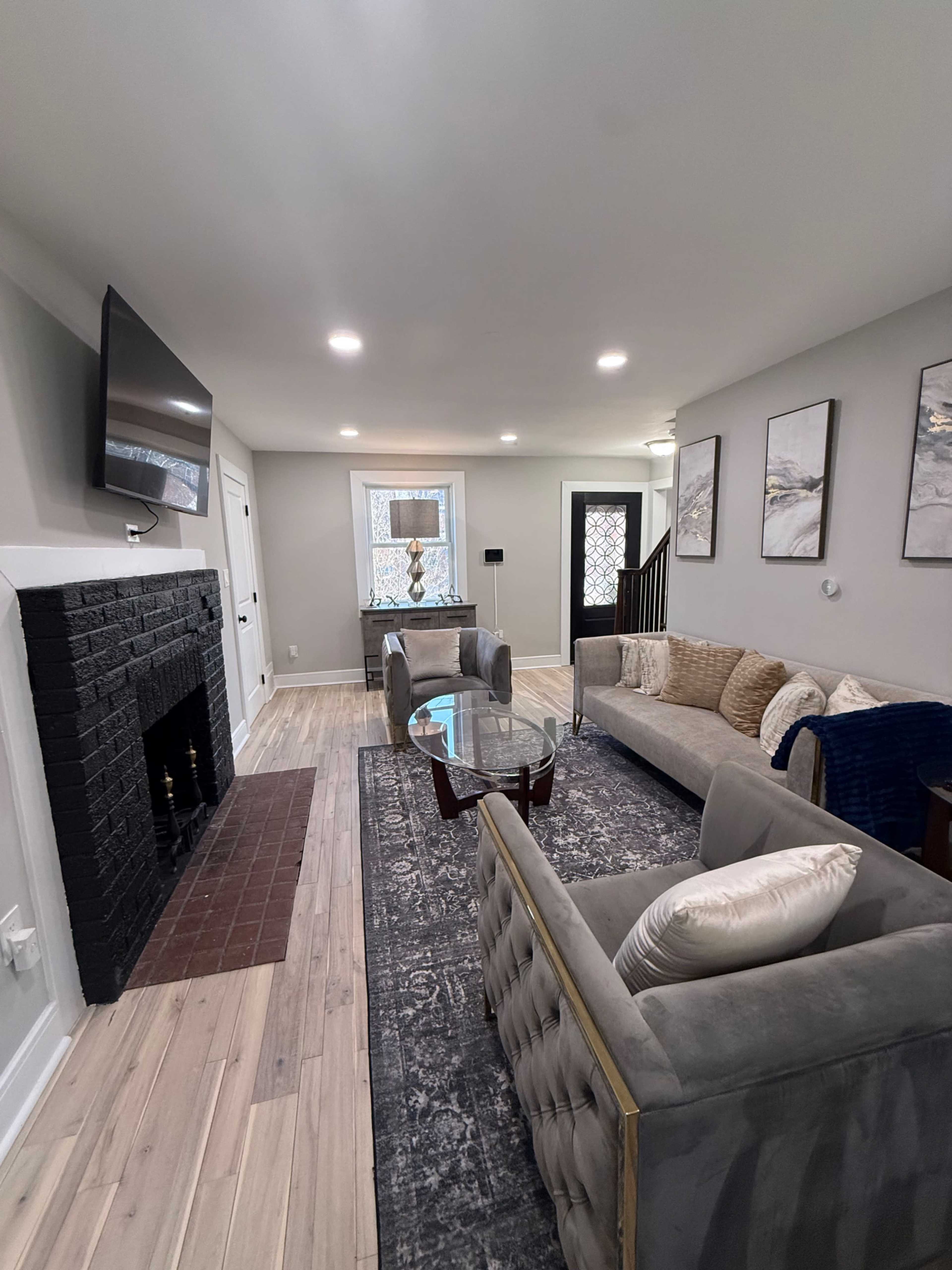 The image shows a modern living room featuring a gray sectional sofa, a glass coffee table, and a black fireplace, with a large window allowing natural light.